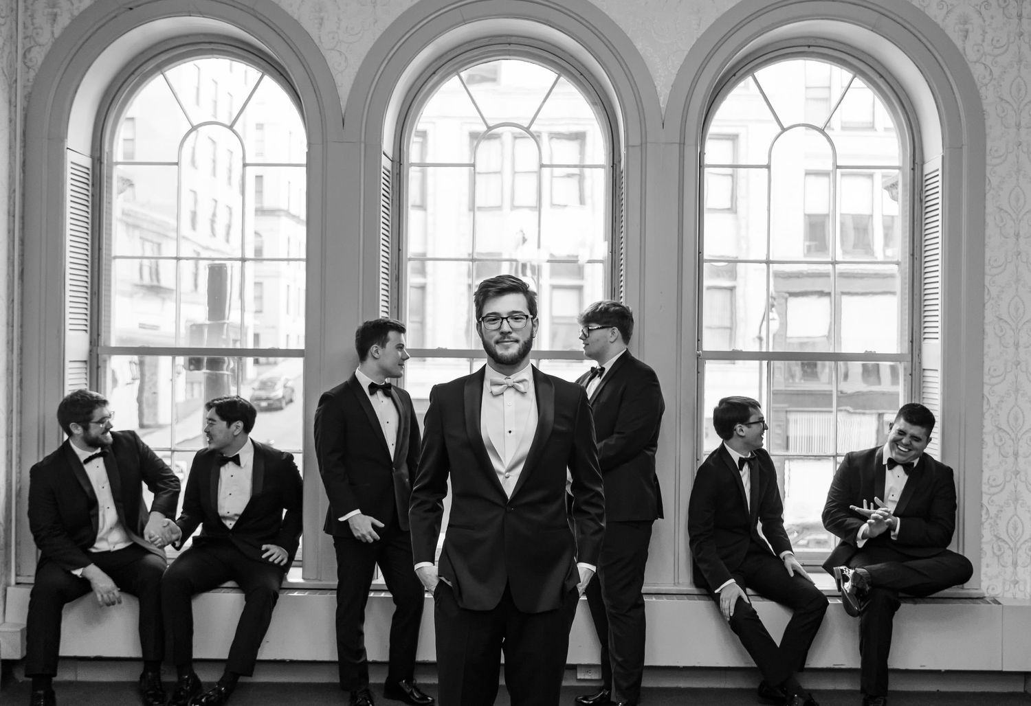 Group in formal attire poses by arched windows in elegant black and white photograph.
