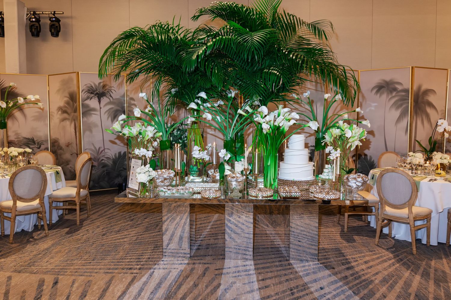 Elegant wedding reception table with towering tropical palm centerpiece, white floral arrangements, and tiered cake in a ballroom.