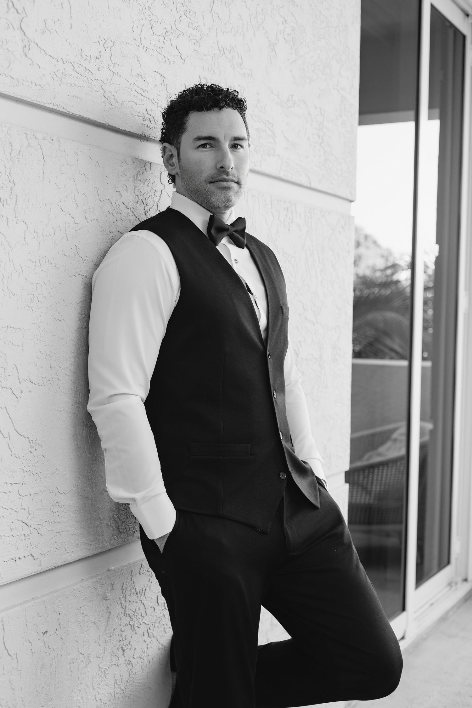 Black and white portrait of a stylish man in a vest and bow tie leaning against a textured white wall.