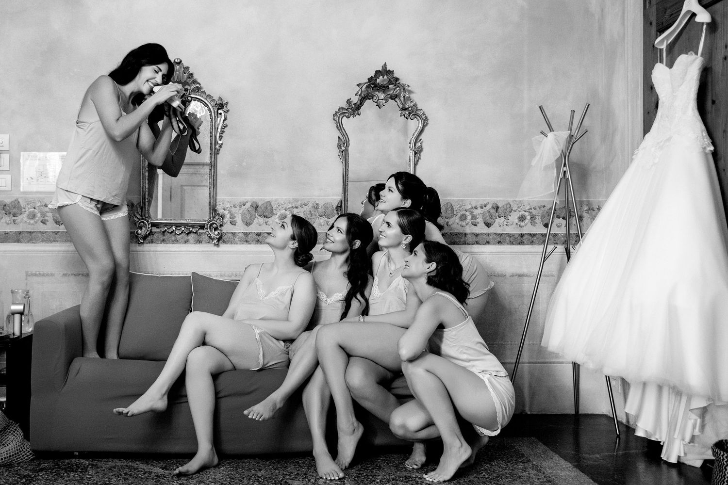 Black and white photo of a photographer capturing a candid moment with bridesmaids in a bridal suite with a wedding dress hanging.