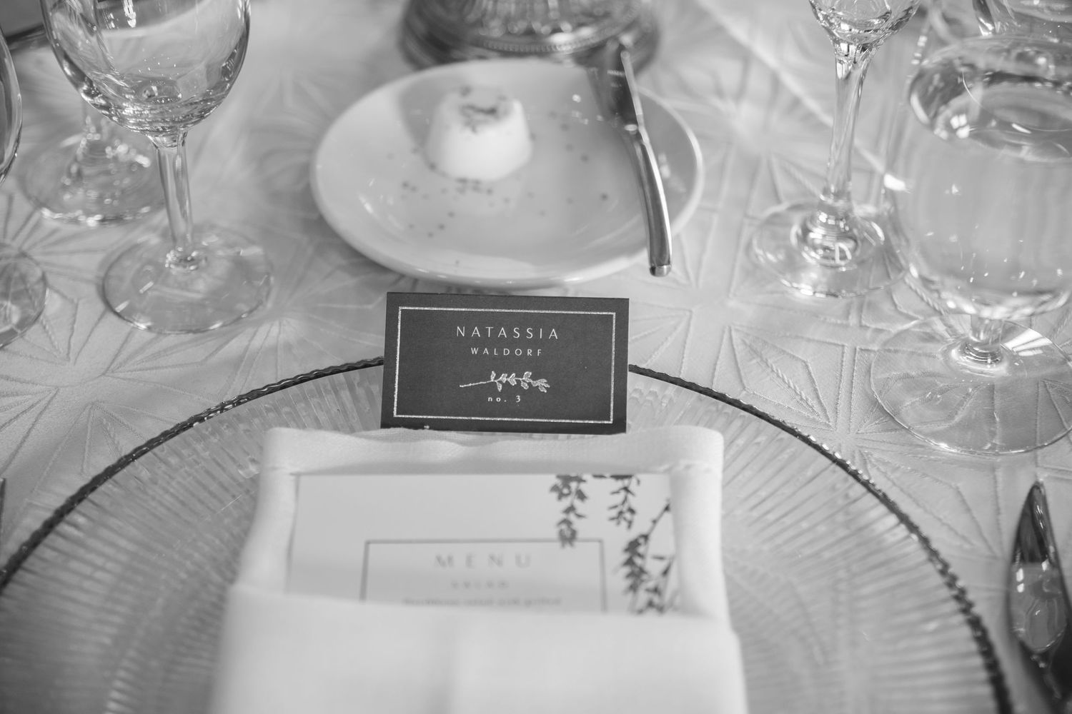 Black and white photo of an elegant wedding place setting with a name card, folded napkin, and crystal glassware.