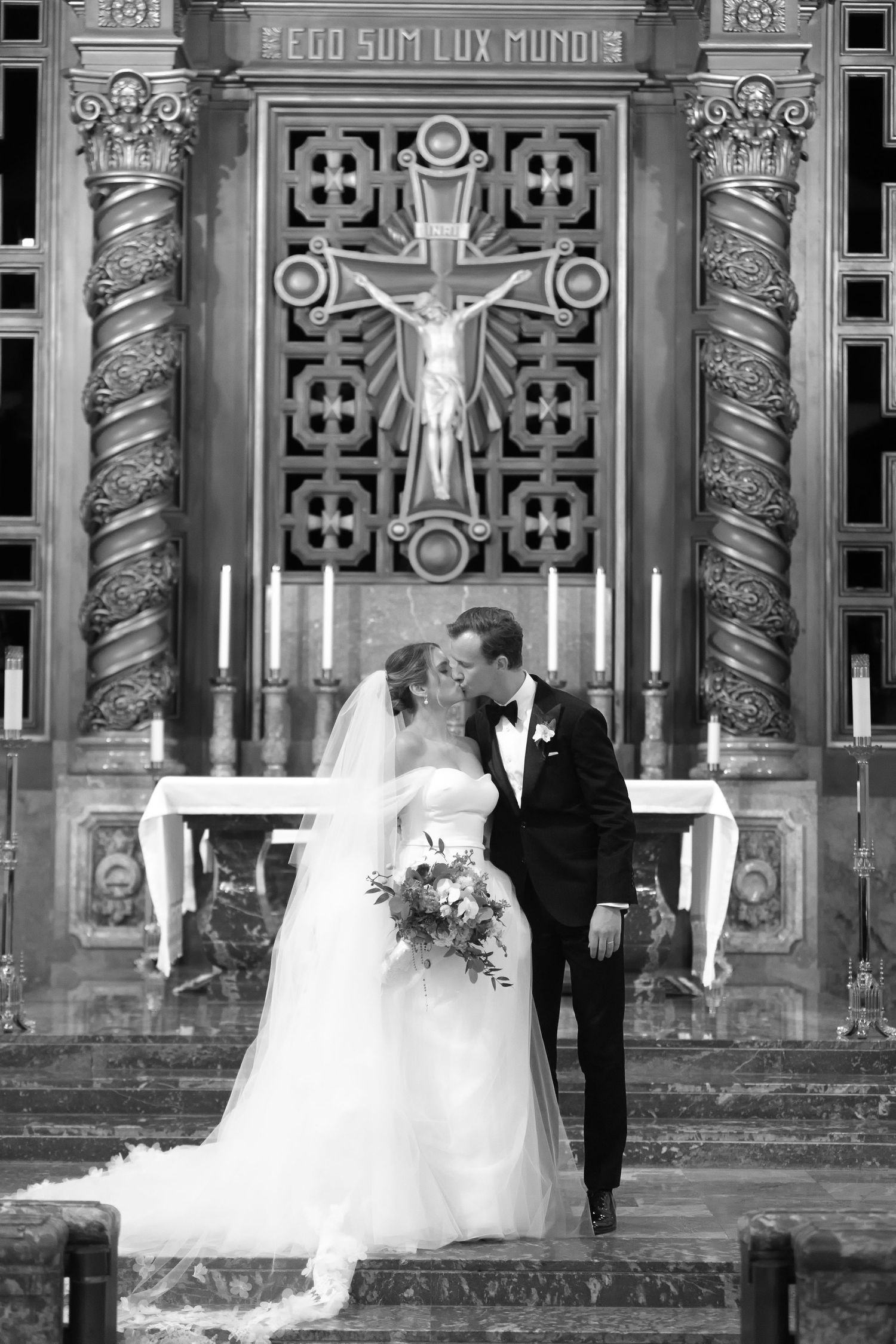 Black and white photo of a bride and groom kissing at the altar of an ornate church with a crucifix backdrop.