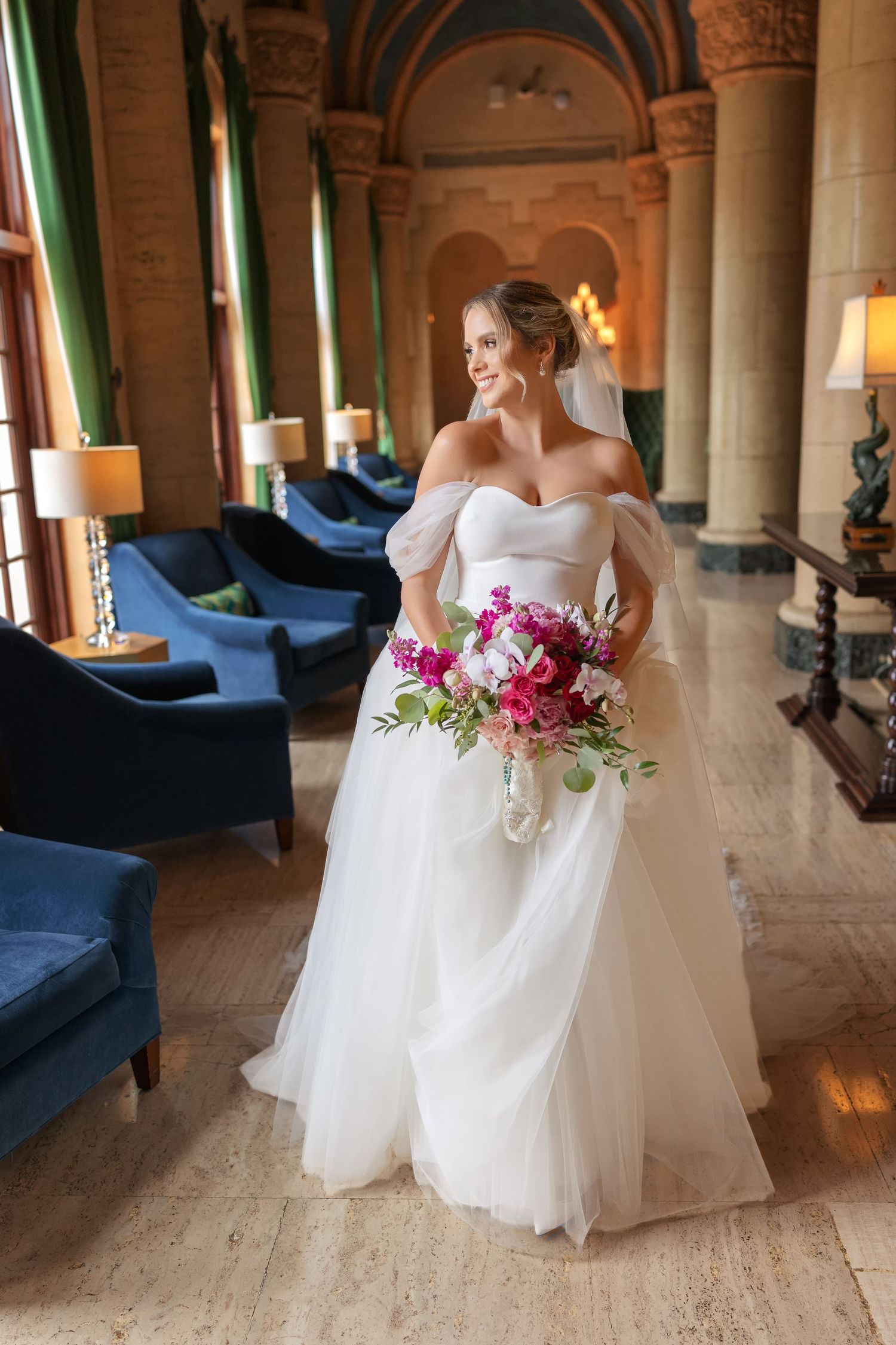 Bride in off-shoulder white tulle gown holds pink bouquet, smiling in elegant venue with blue velvet chairs.
