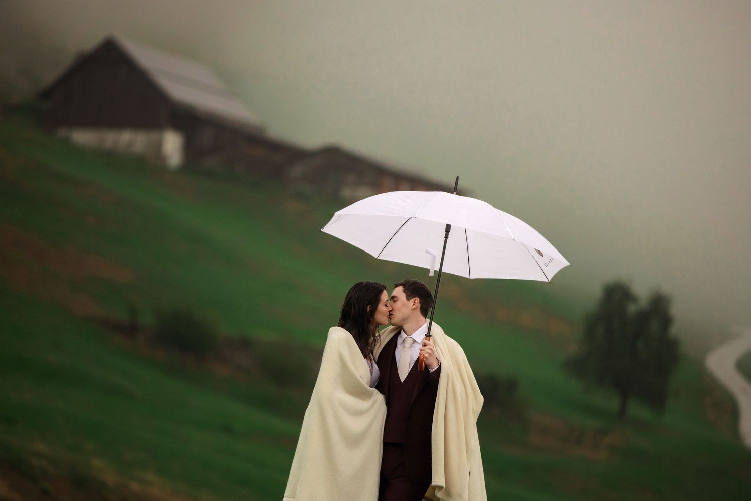 Couple kissing under white umbrella on misty green hillside with barn in background.