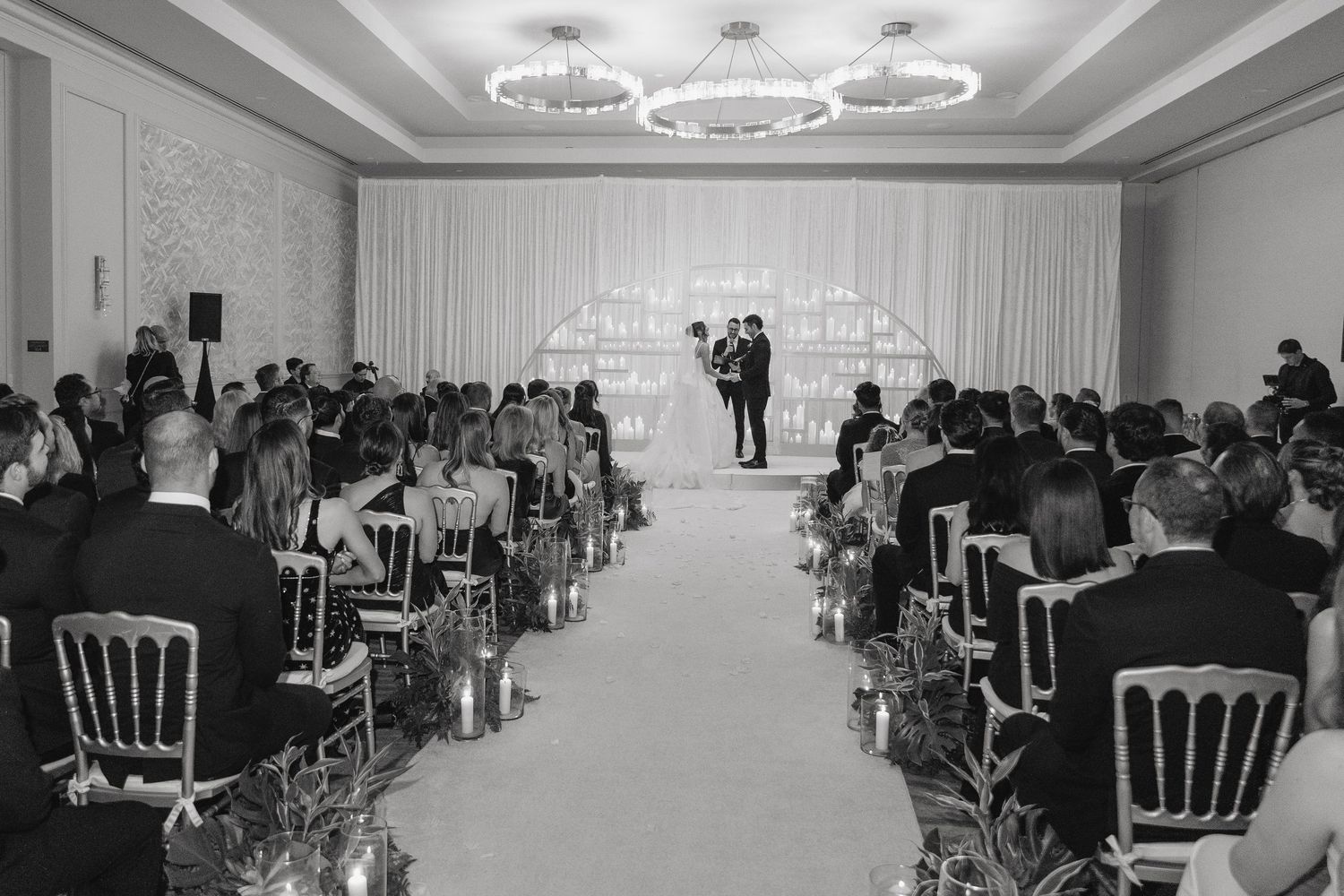 Black and white photo of an elegant indoor wedding ceremony with guests seated in rows facing the couple at a decorated altar.