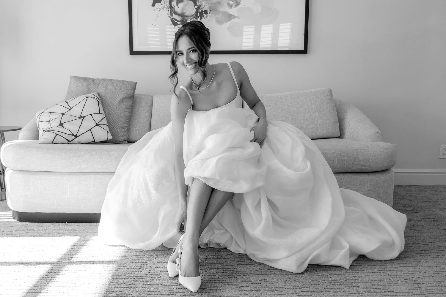 Bride in white ball gown sitting on sofa, adjusting heels, black and white wedding portrait.