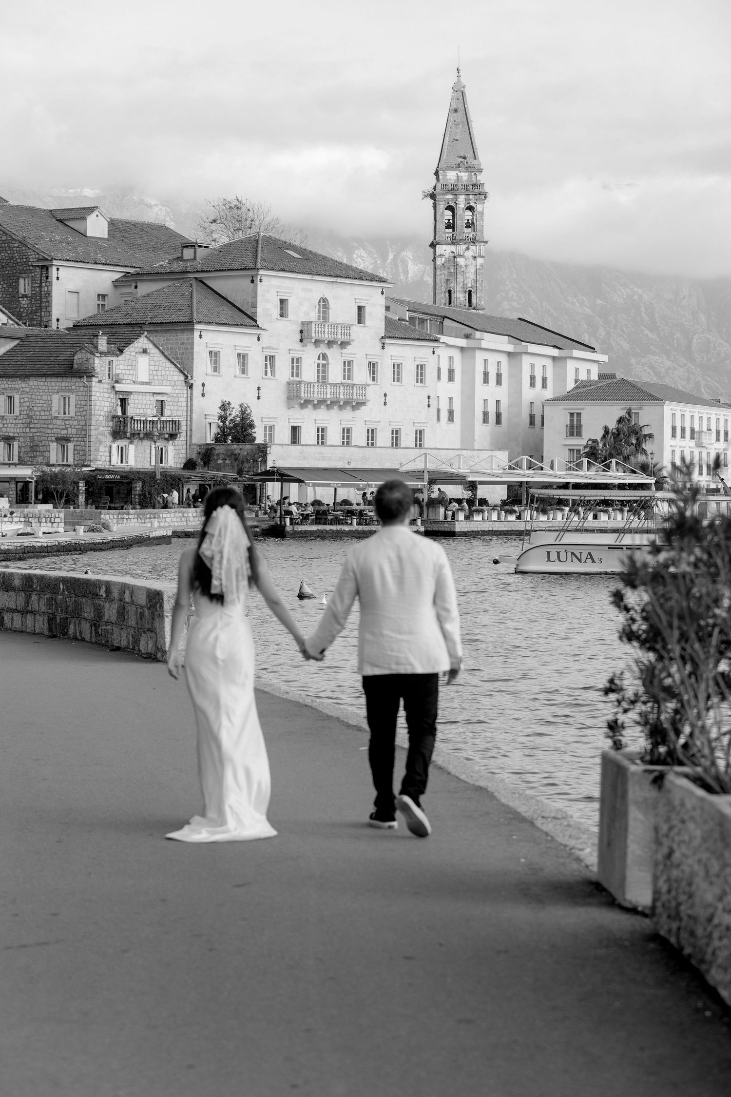 A couple walks along a waterfront promenade with historic buildings and a church bell tower in the background.