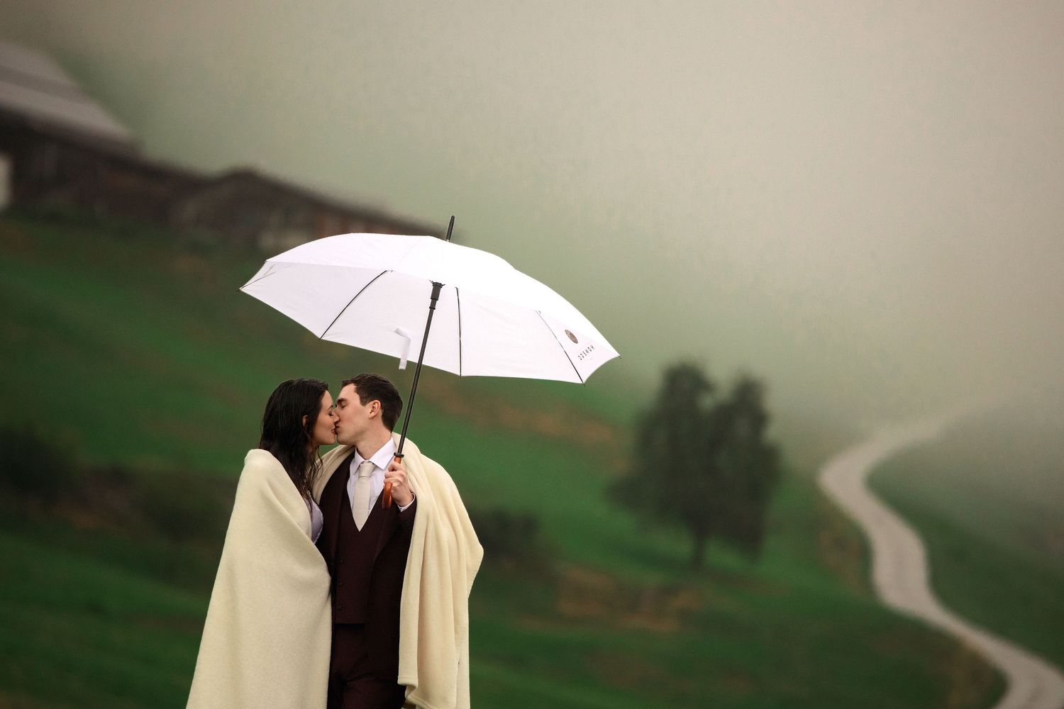 Couple kissing under a white umbrella in the rain with lush green hills and a winding road in the background.