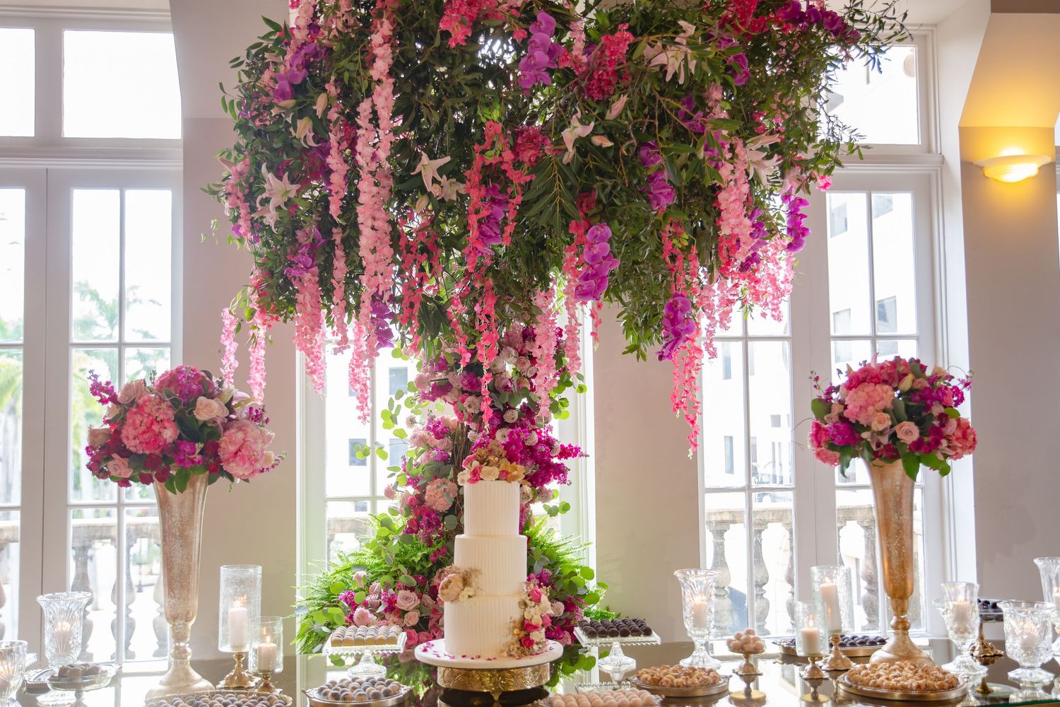 Elegant wedding dessert table with white cake under a lush floral chandelier of pink and purple blooms in a bright venue.