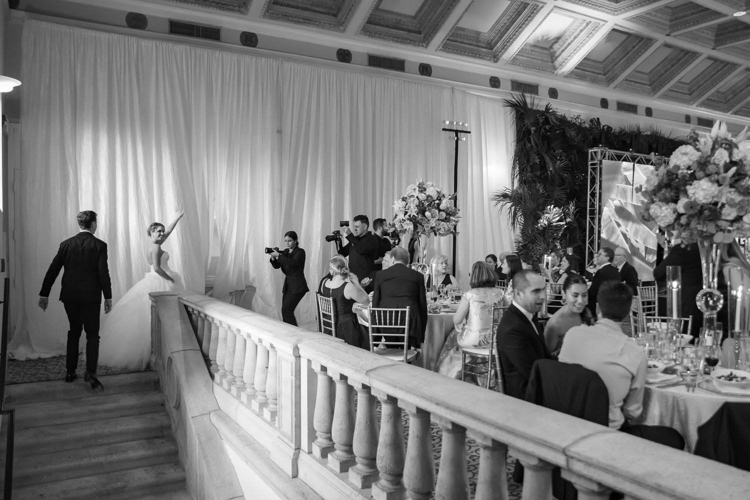 Black and white photo of bride and groom making grand entrance at elegant wedding reception with guests seated at decorated tables.