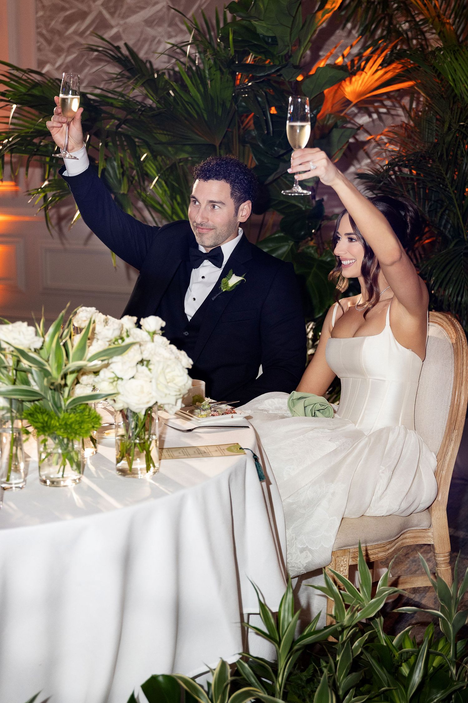 A bride and groom toast champagne at their wedding reception table decorated with white floral centerpieces.