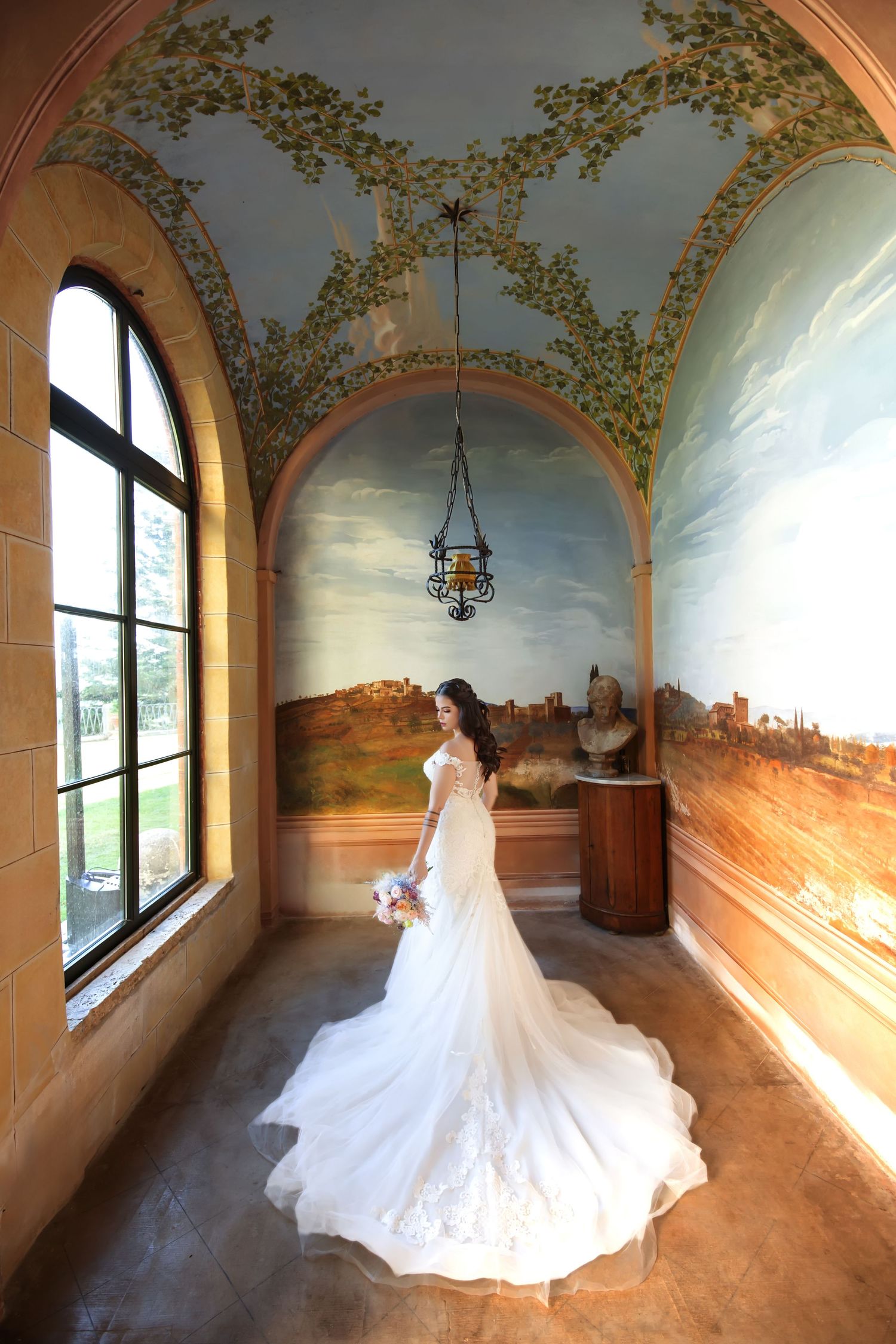 Wedding dress with long train displayed in ornate hallway with painted murals and arched windows.