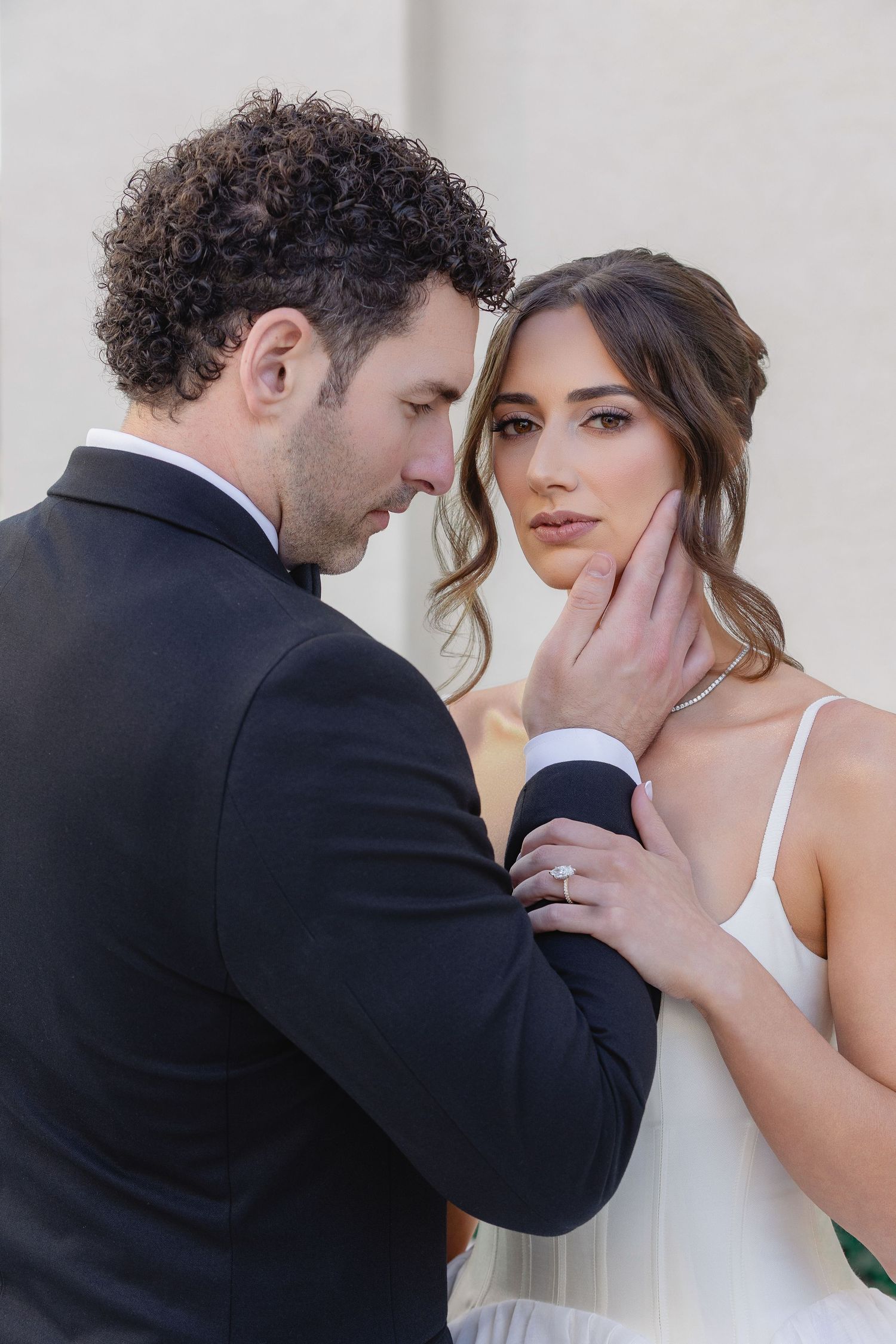 Groom in black suit tenderly holds bride's face as she gazes forward in white dress, wearing engagement ring.