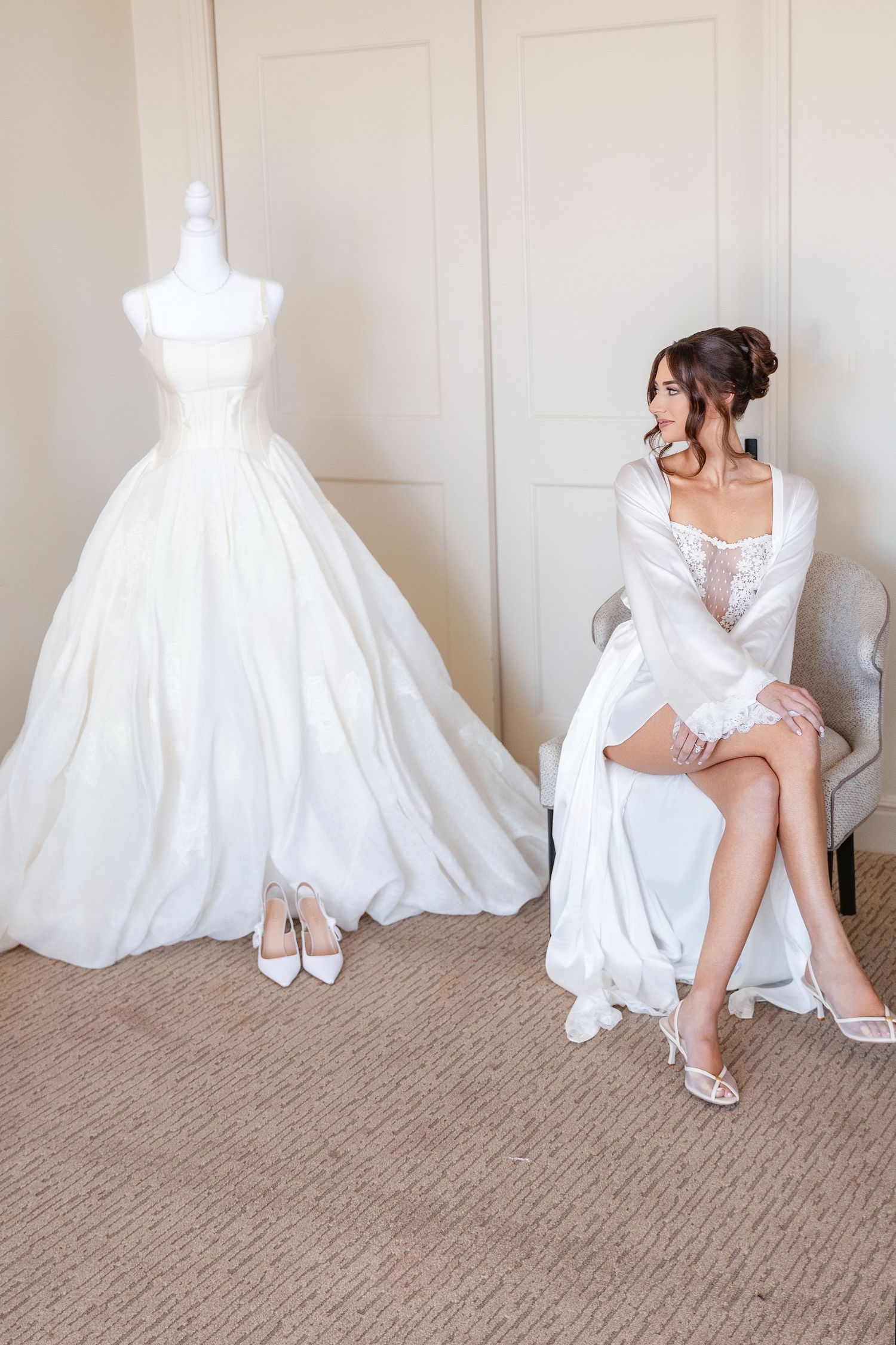 Bride in white robe sits beside elegant ball gown on mannequin with heels on floor in bright bridal suite.