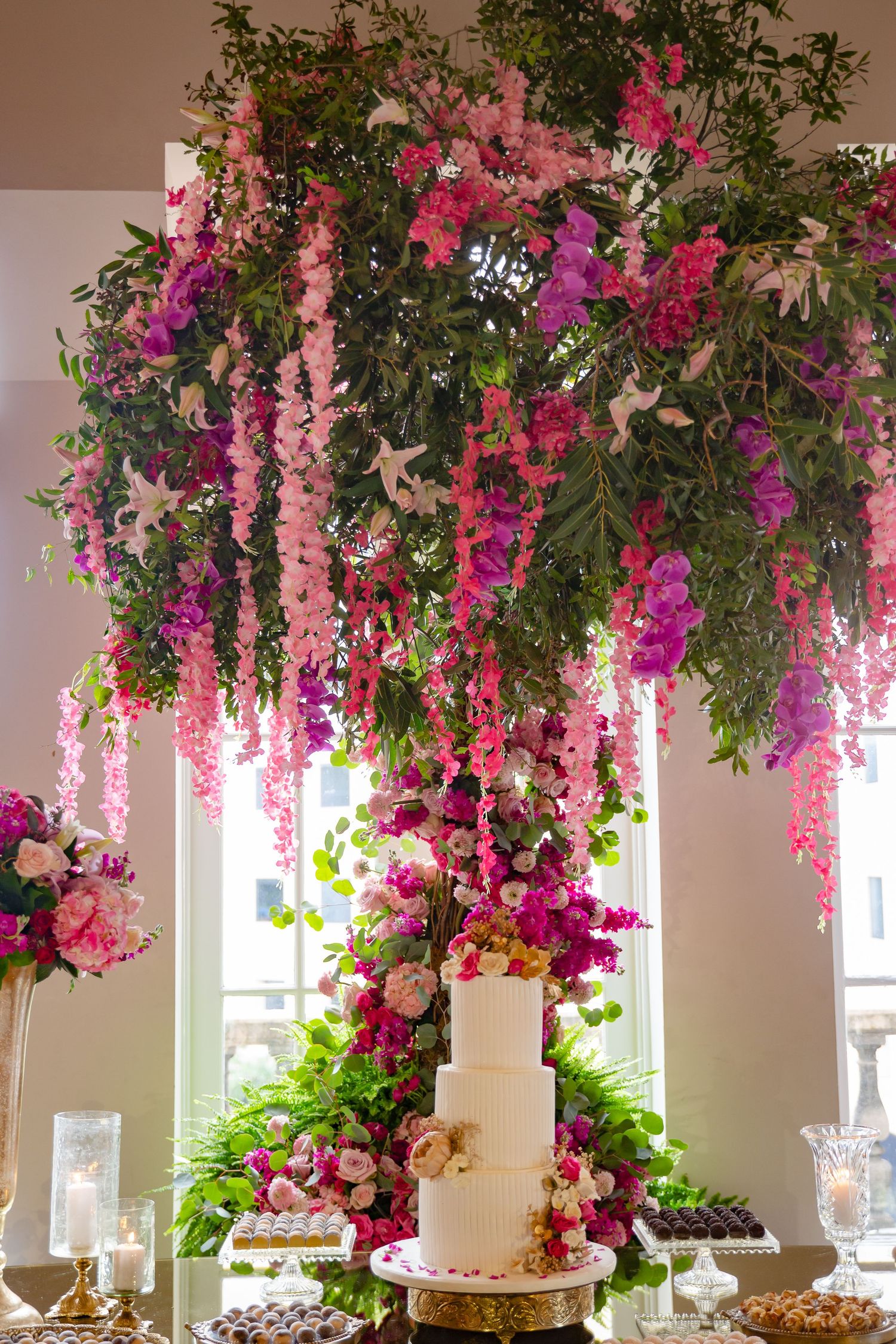 Elegant wedding cake display under cascading pink and purple floral installation with greenery in a bright venue.