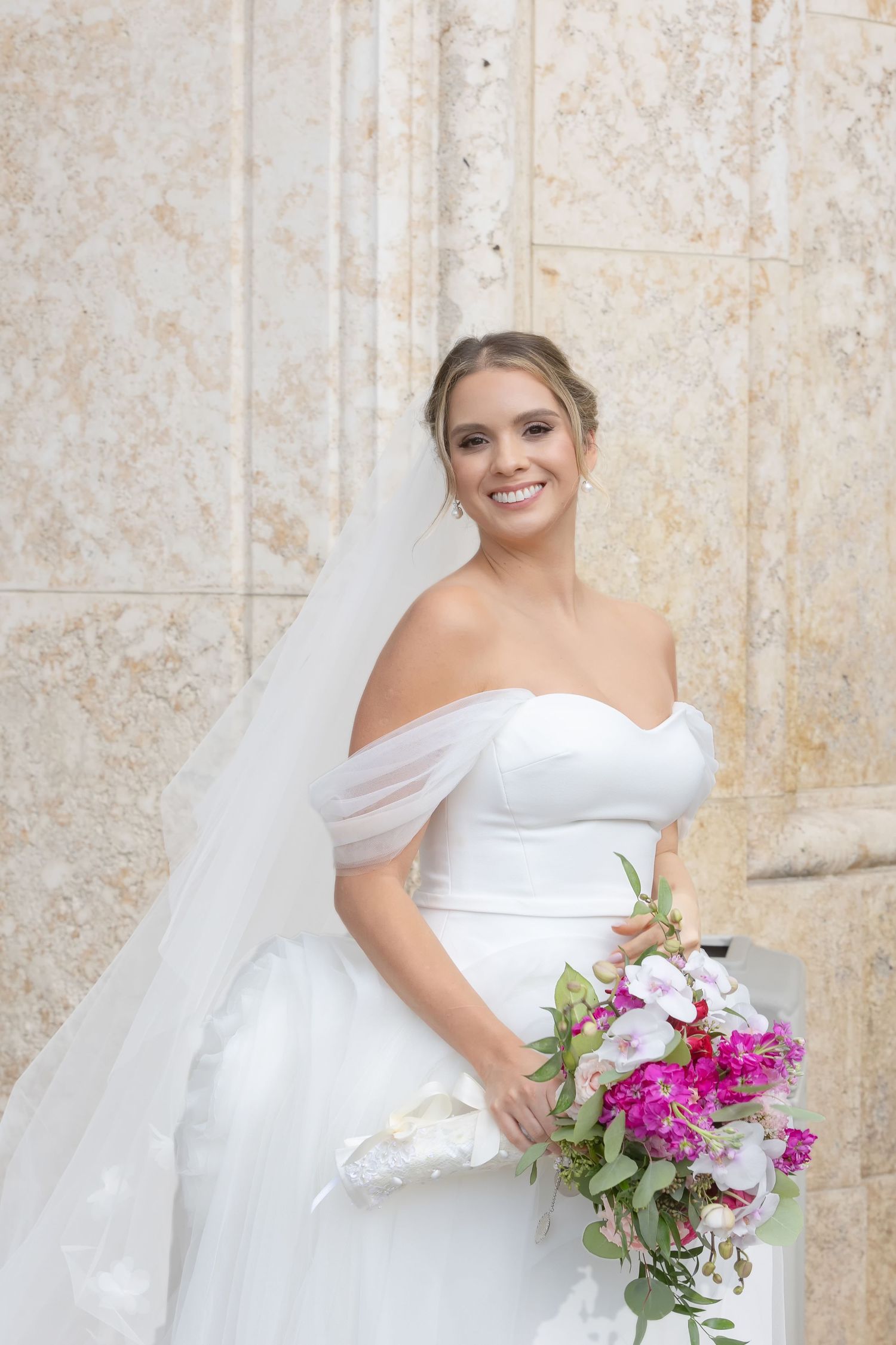 Smiling bride in white off-shoulder gown and veil holds pink and white bouquet against marble wall.