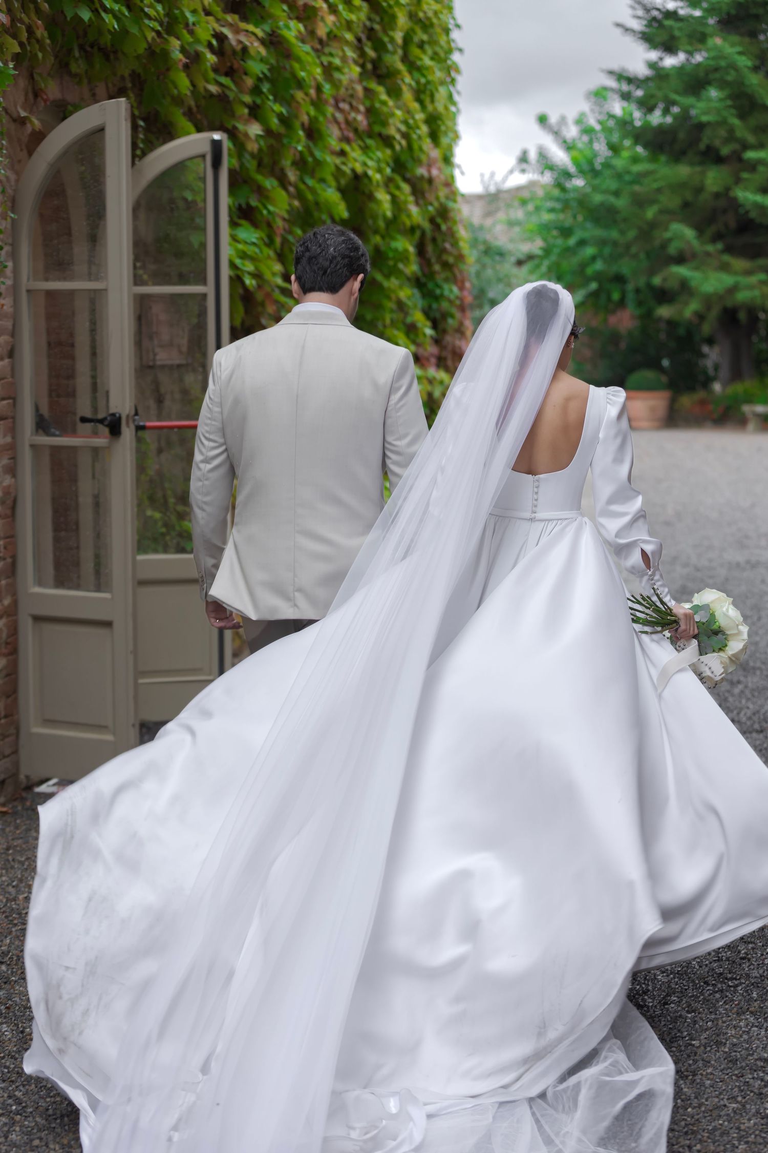 A bride in a flowing white gown and veil stands behind a groom in a light gray suit near a vine-covered doorway.