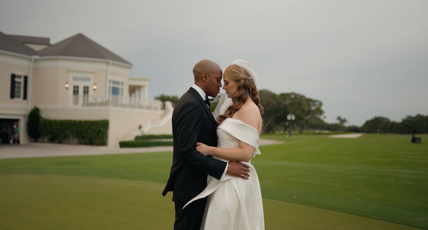 A couple in formal wedding attire embraces while standing on a lush green golf course at sunset.