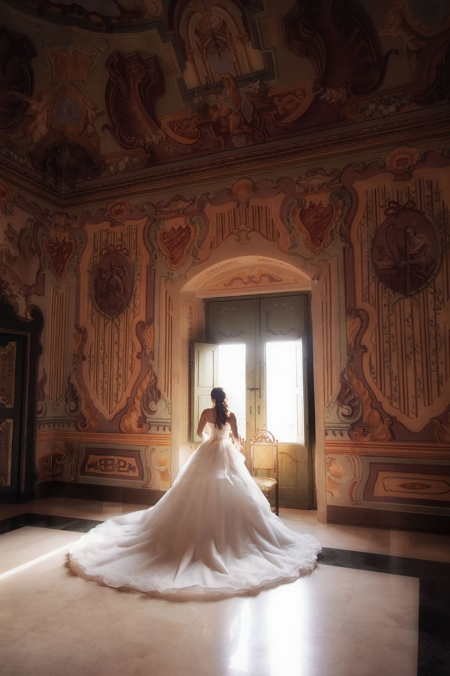 Bride in white ball gown with long train stands before ornate baroque window in elegant frescoed Italian villa.