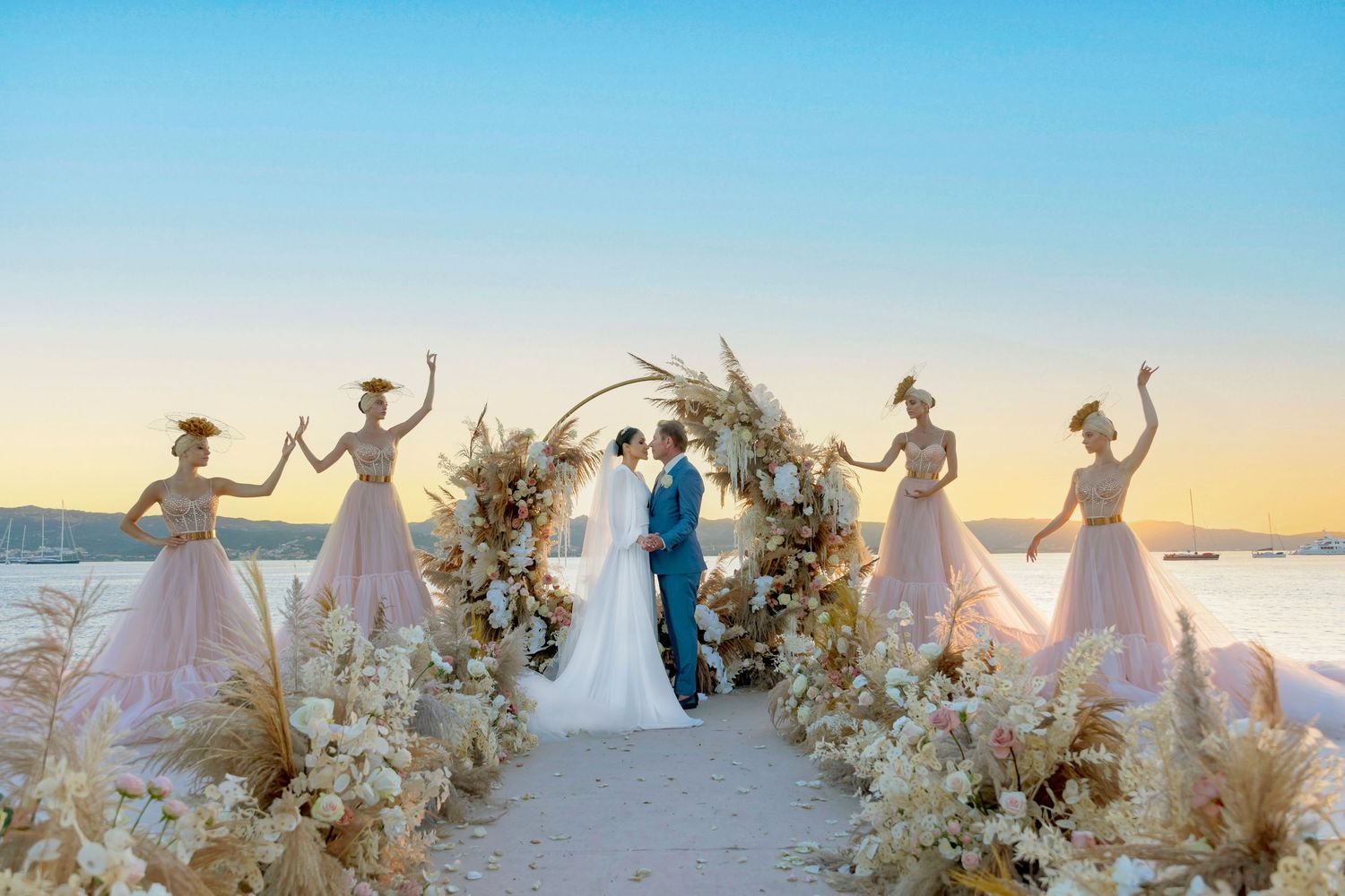 Romantic beach wedding ceremony under a floral arch with attendants in pink dresses at sunset.