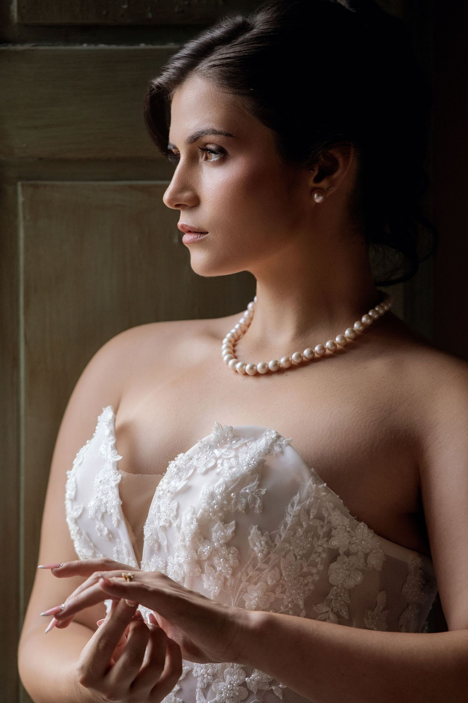 Elegant bride in lace strapless gown wearing pearl necklace and earrings, gazing thoughtfully in soft dramatic lighting.