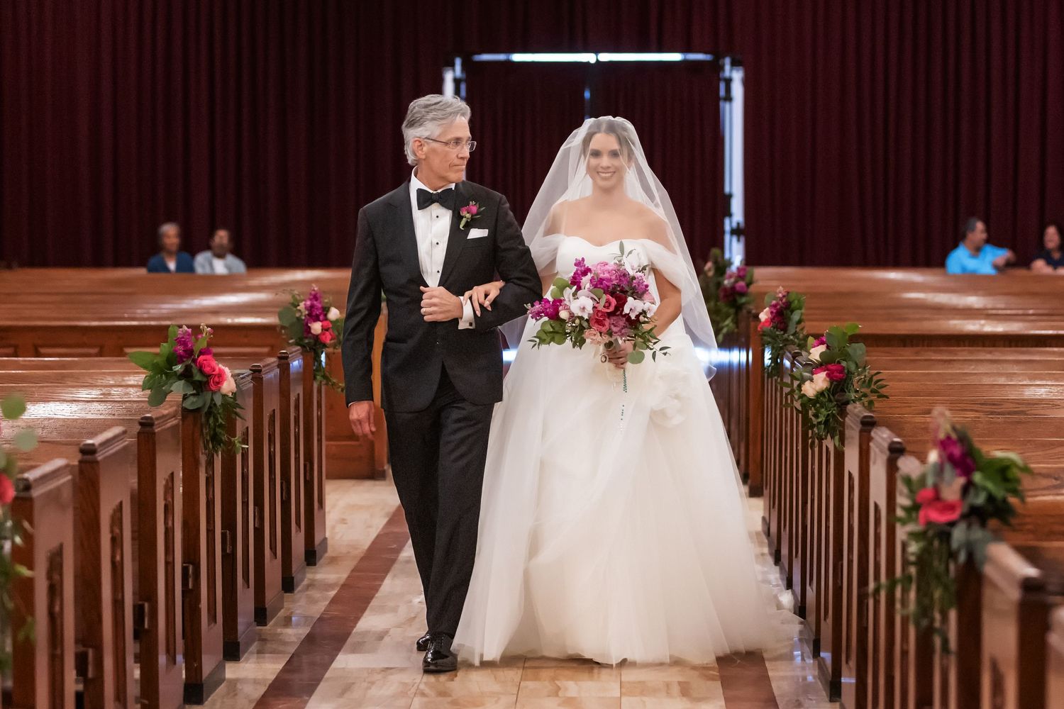 Bride in white ball gown walks down church aisle with father in tuxedo, flanked by floral-adorned wooden pews.