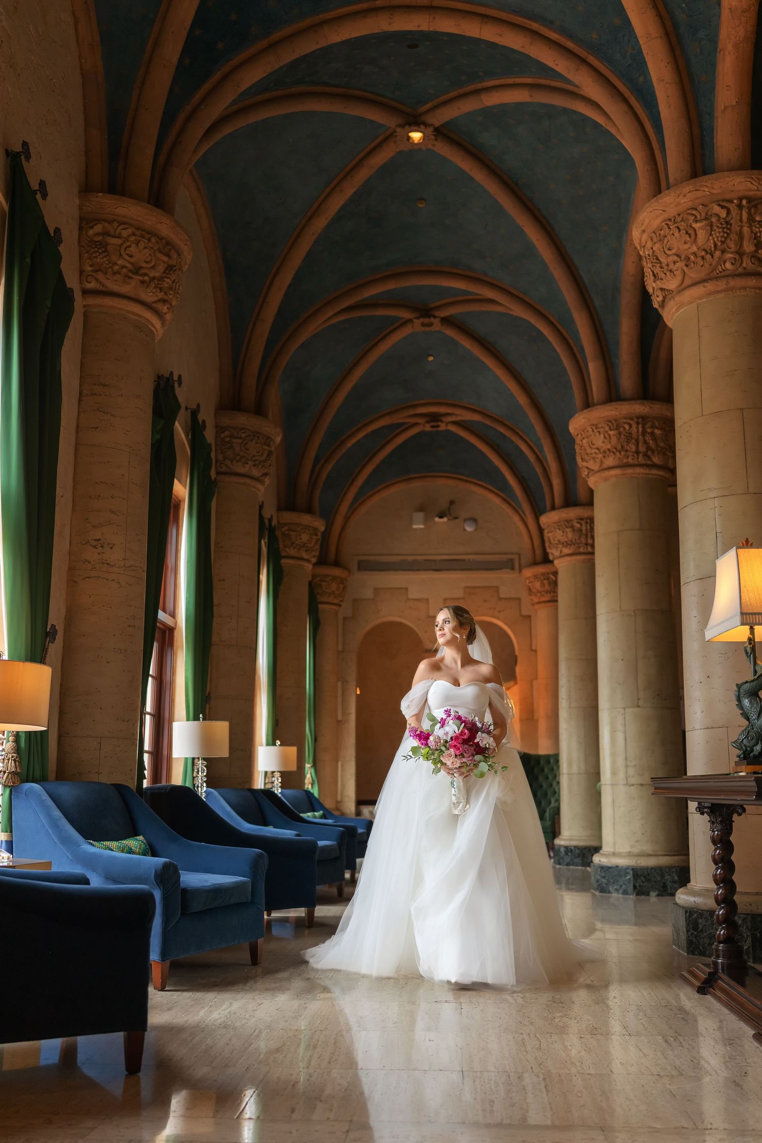 Bride in white gown holding pink bouquet stands in elegant vaulted hall with ornate columns and blue velvet chairs.