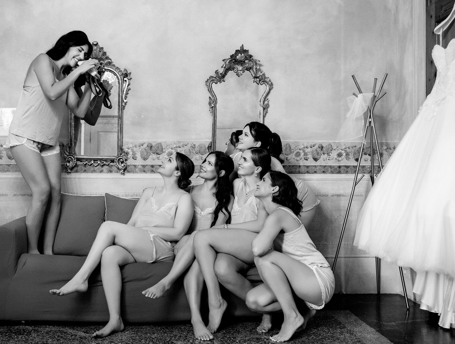 Black and white photo of bridesmaids posing on a sofa while one photographs them, with a wedding dress displayed nearby.