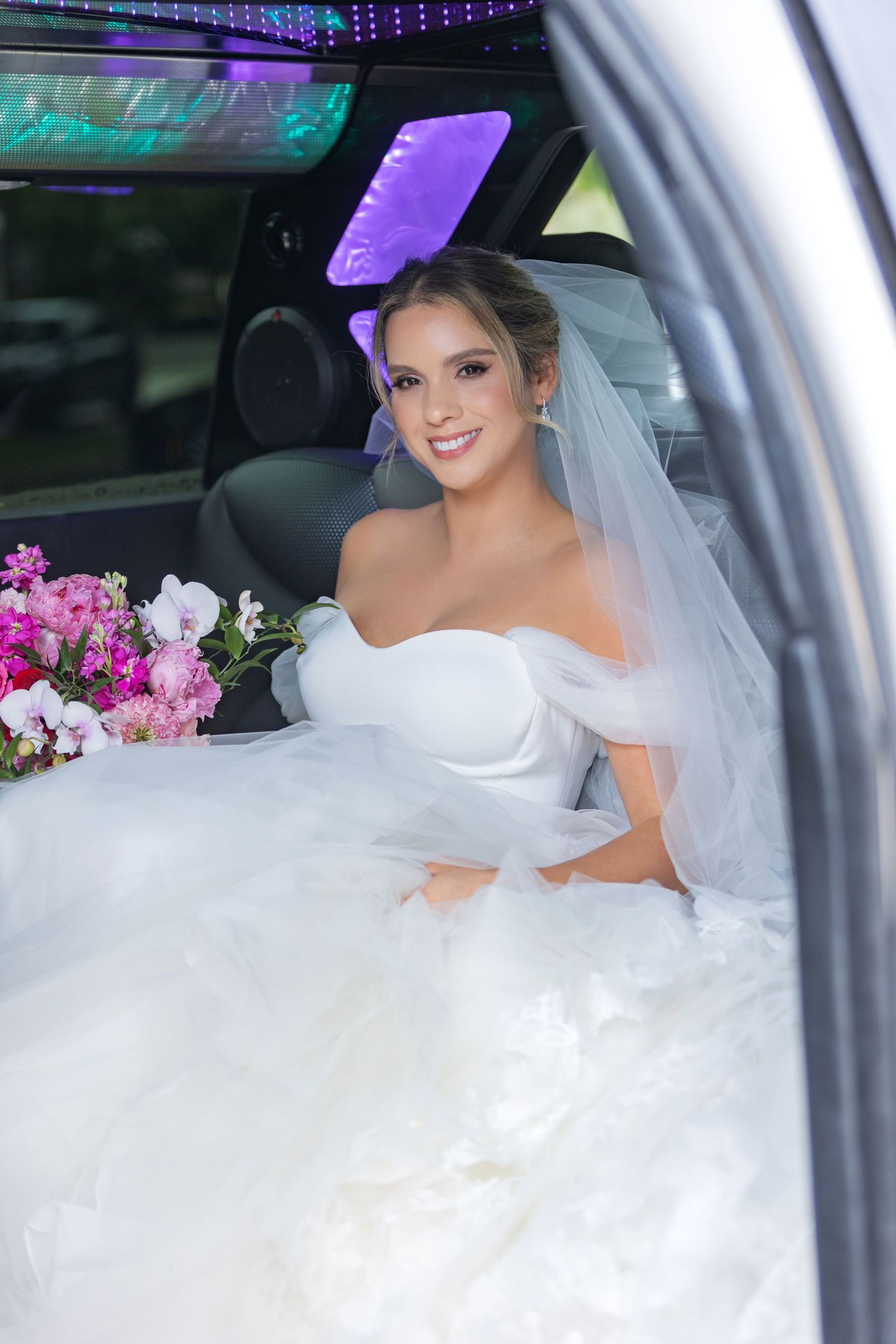 Smiling bride in white off-shoulder gown and veil sits in a decorated limousine holding a pink floral bouquet.