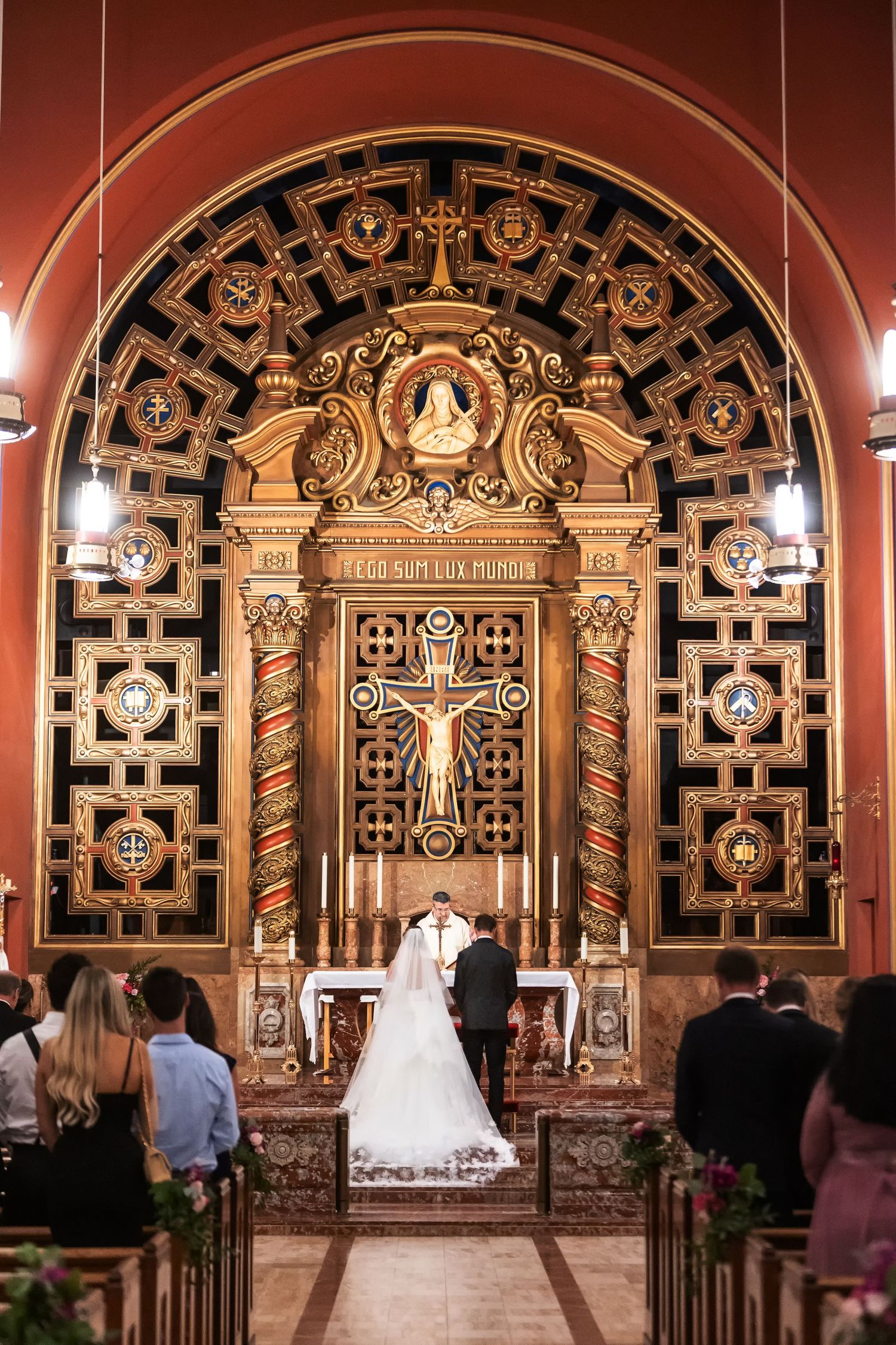 Bride and groom stand at ornate golden altar during Catholic church wedding ceremony with intricate decorative archway.