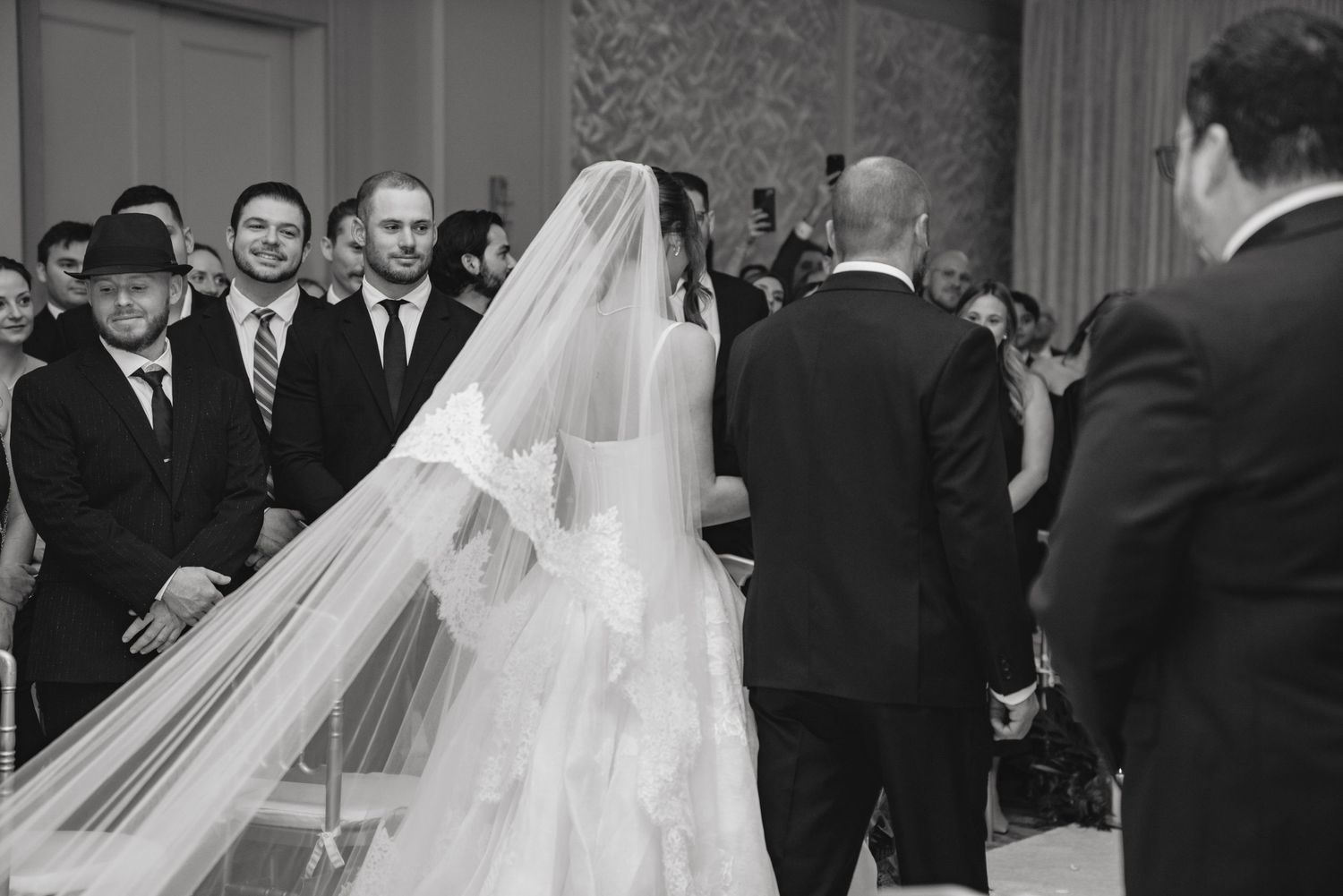 Black and white wedding ceremony photo showing bride in lace gown and veil facing groom before guests.