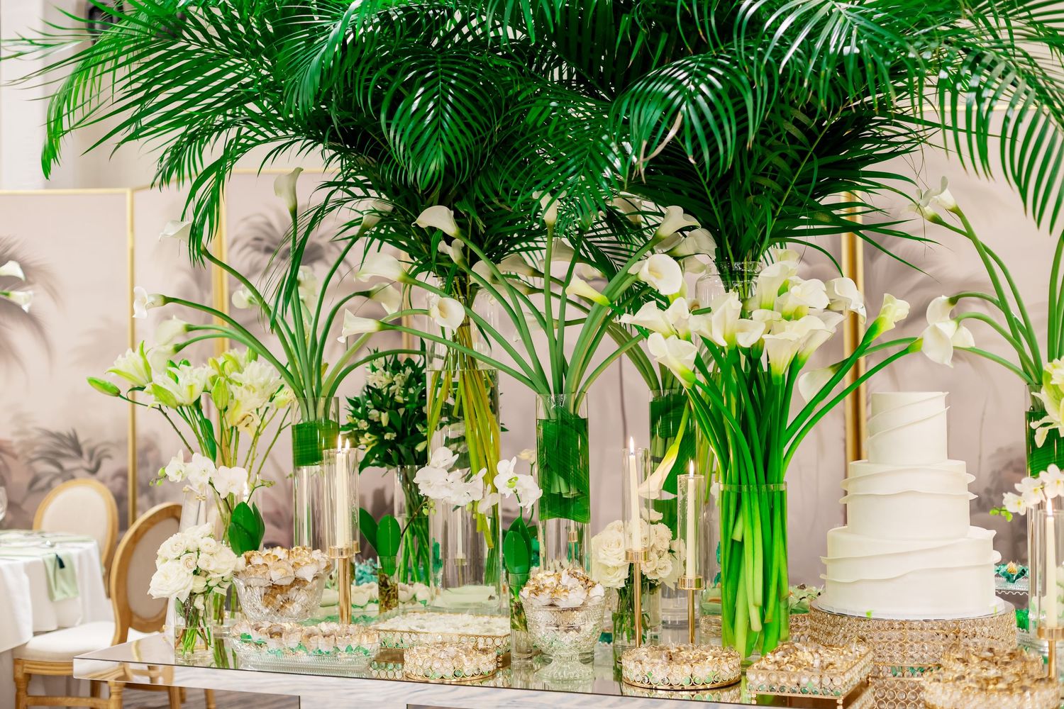 Elegant wedding dessert table with white cake, tropical palm leaves, and lush green and white floral arrangements in glass vases.