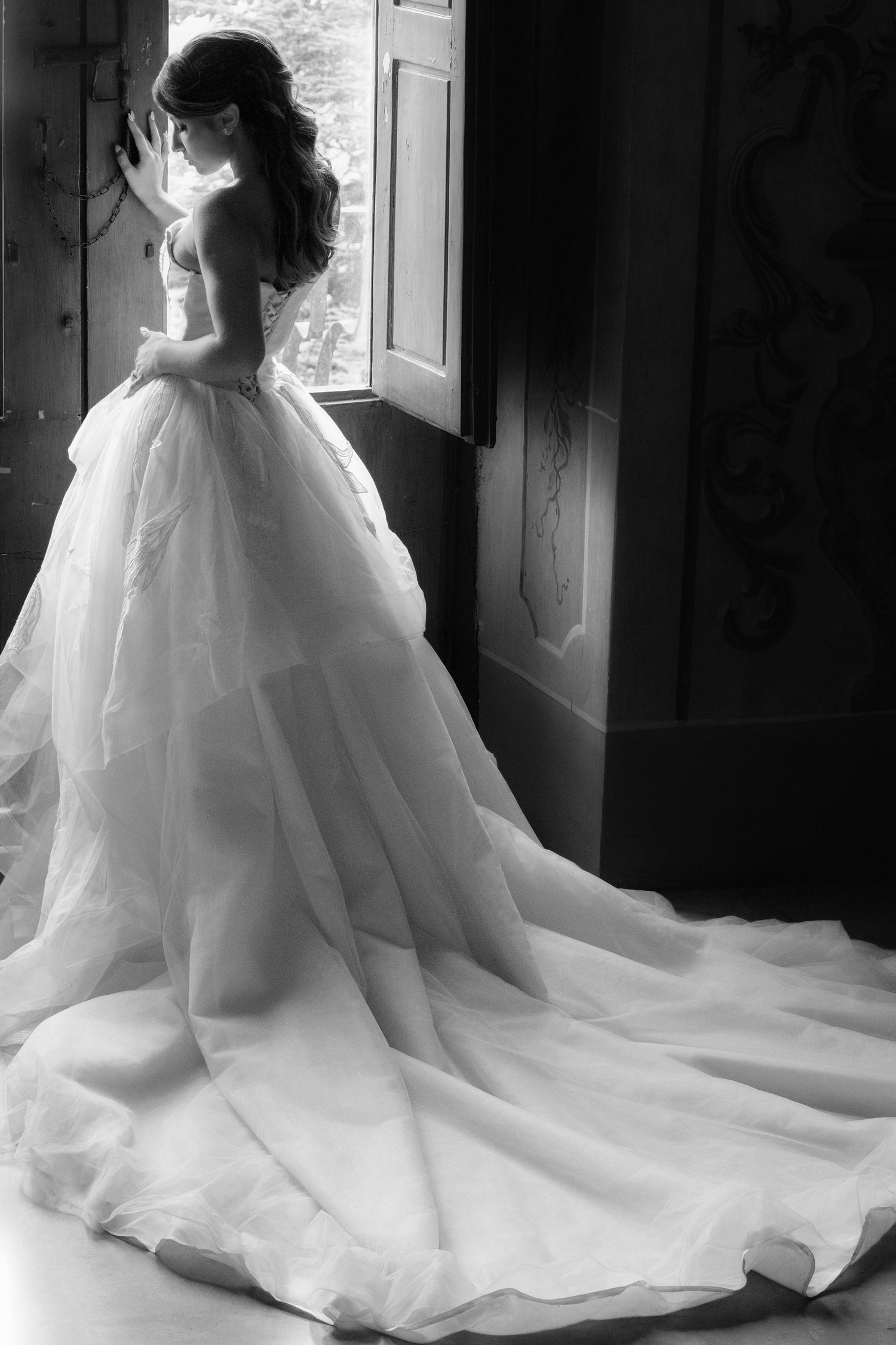 Bride in a stunning ball gown with long train stands by window in black and white wedding photo.