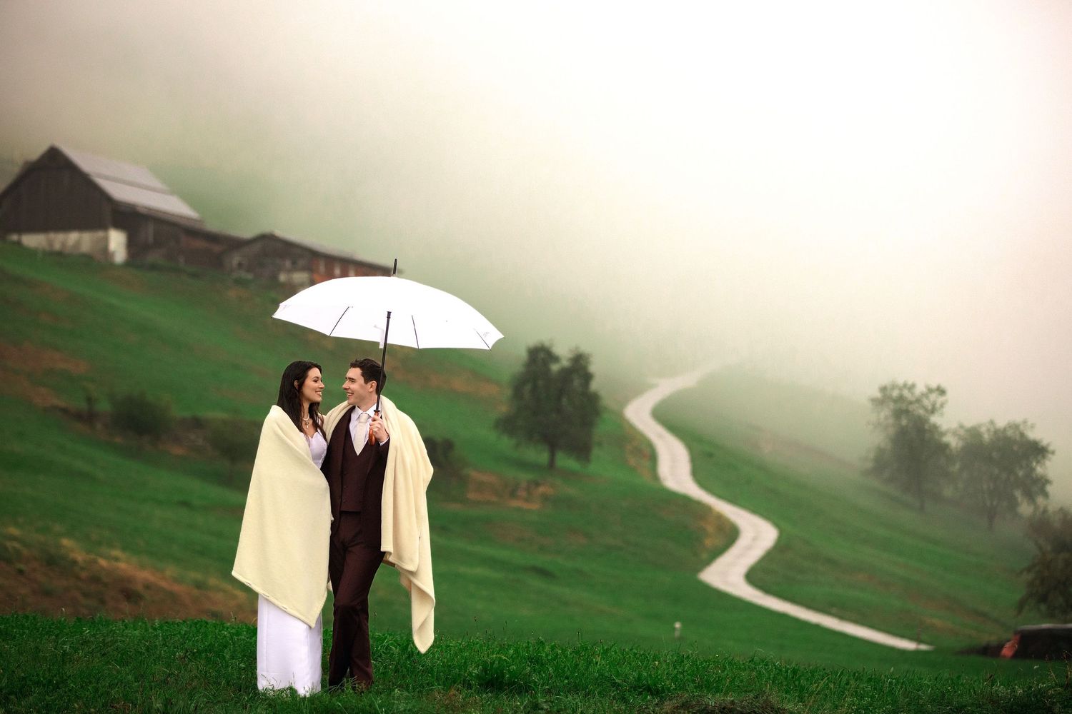 Couple sharing a white umbrella on a misty green hillside with a winding path and barn in the background.