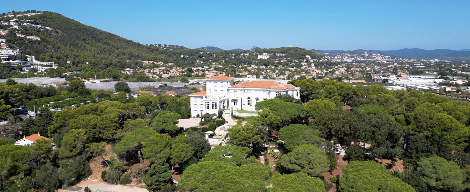Panoramic aerial view of coastal Mediterranean town with mountains, white buildings, and lush green landscape.