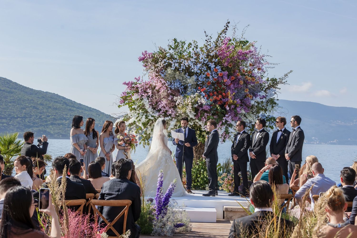 Outdoor wedding ceremony overlooking a scenic mountain lake with purple floral arch decorations and guests seated.