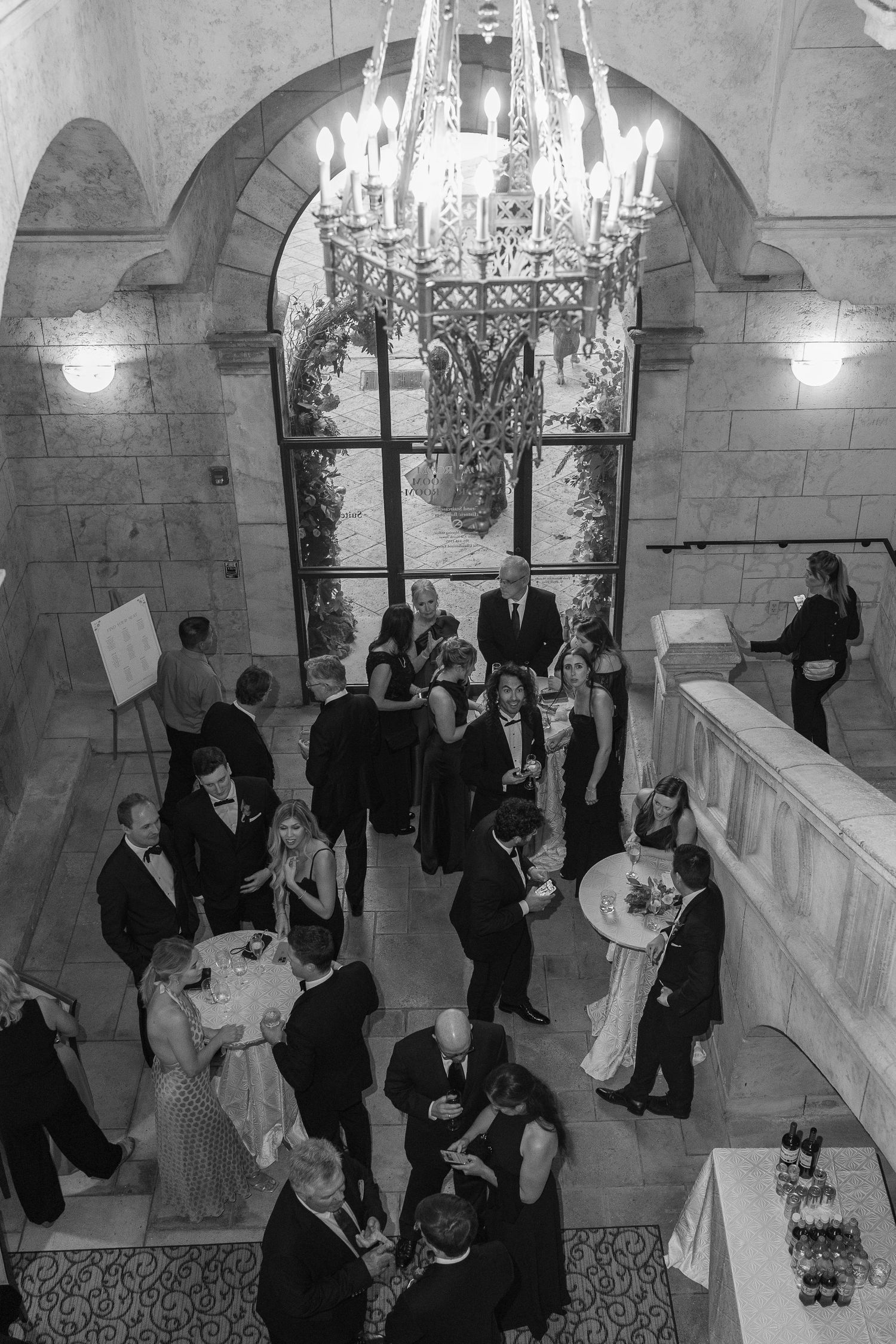 Black and white aerial view of elegantly dressed guests mingling at a formal gala beneath a grand chandelier.