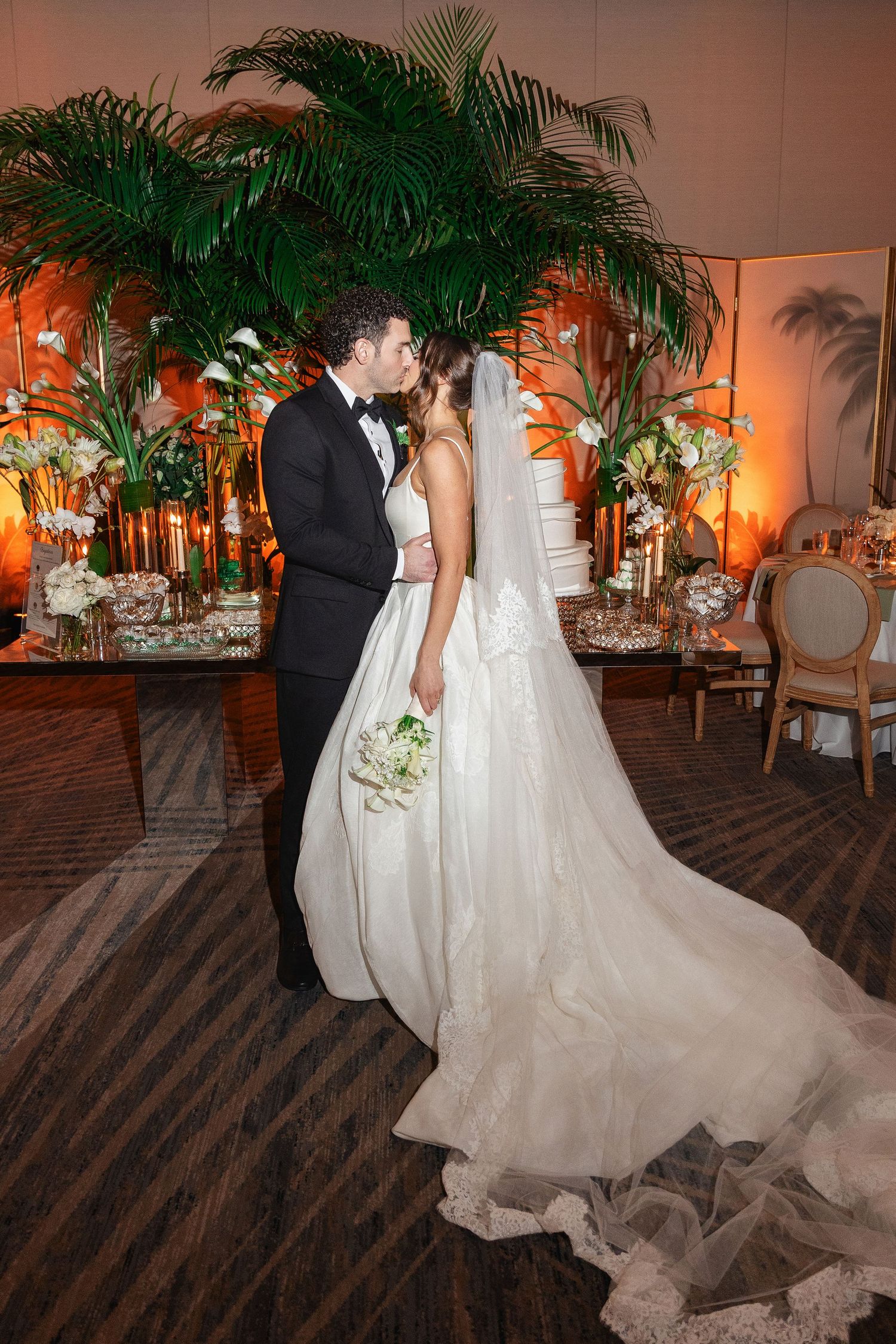 Bride and groom share a kiss in elegant wedding venue with tropical floral arrangements and warm amber lighting.