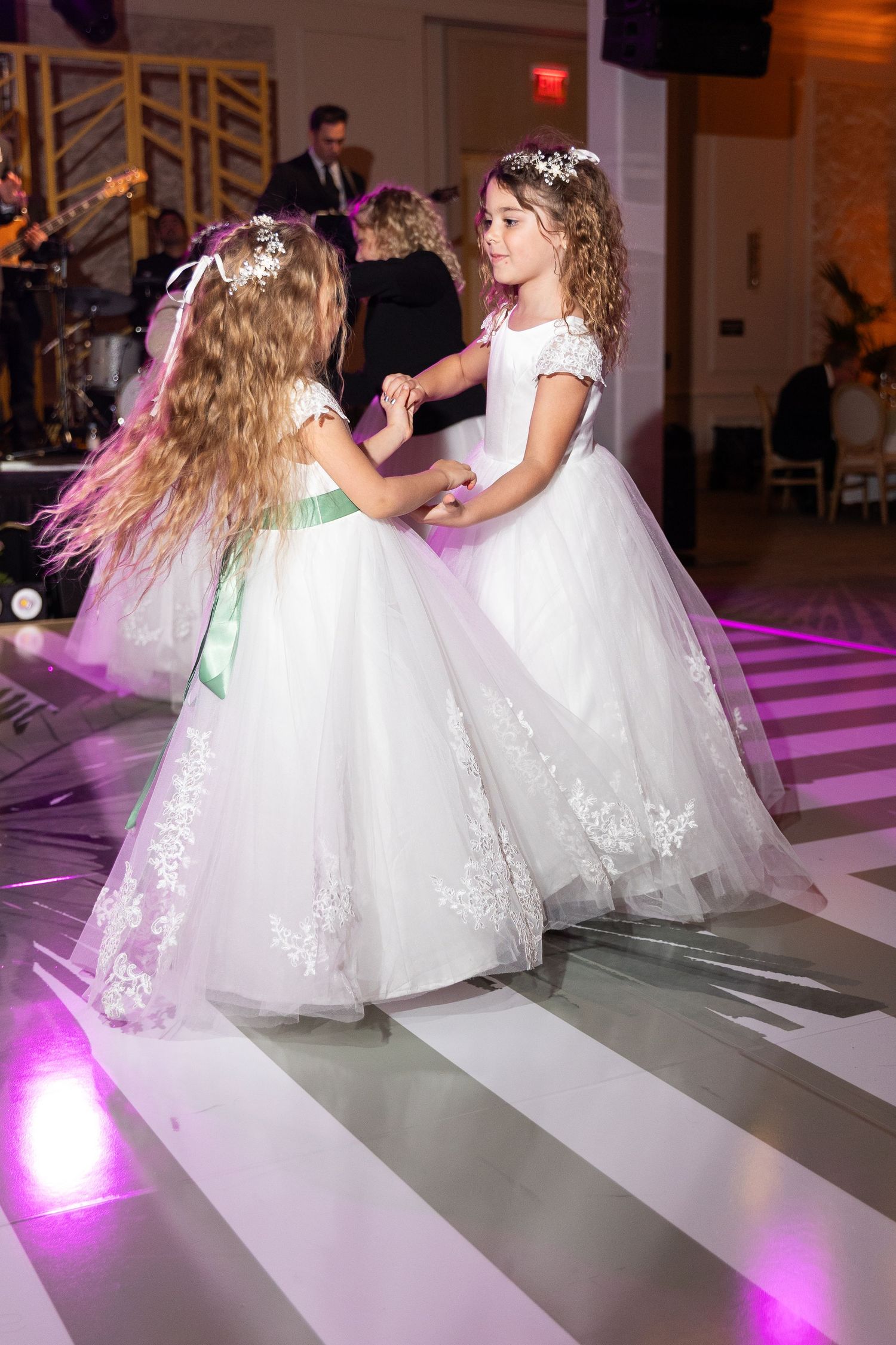 Two flower girls in white lace ball gowns dance together on a striped dance floor at a wedding reception.
