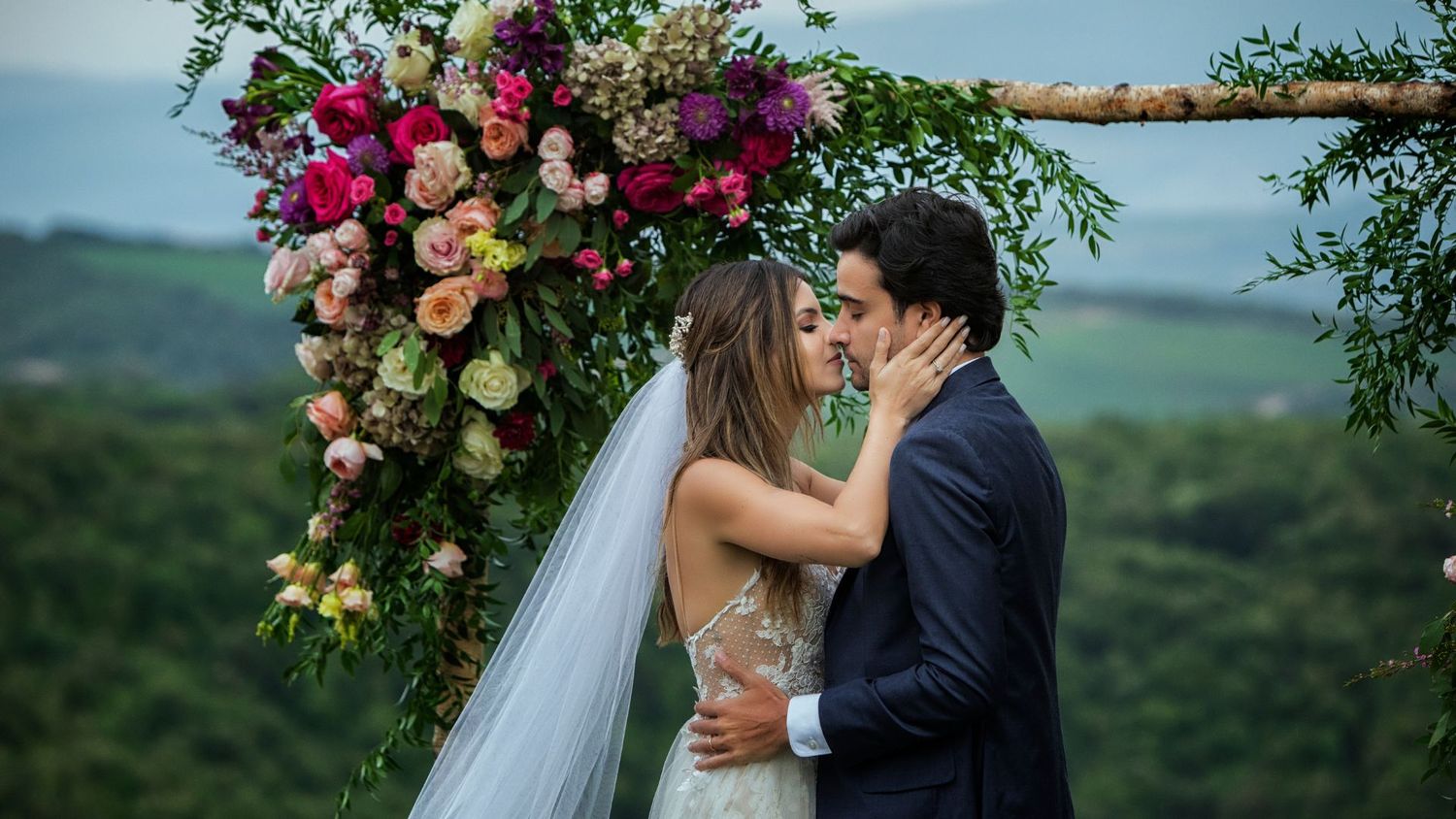 Wedding ceremony under floral arch with vibrant roses and mountain landscape backdrop.