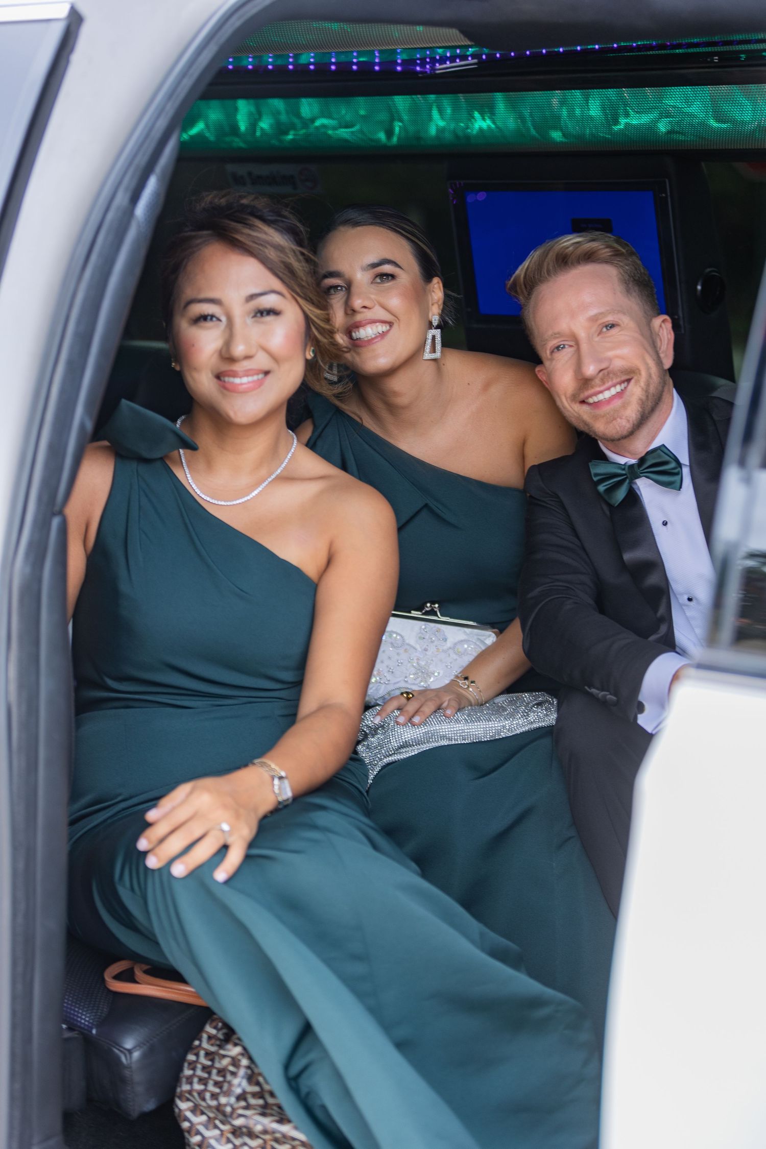 Three elegantly dressed friends smile in a limousine, wearing matching dark green formal attire for a special event.