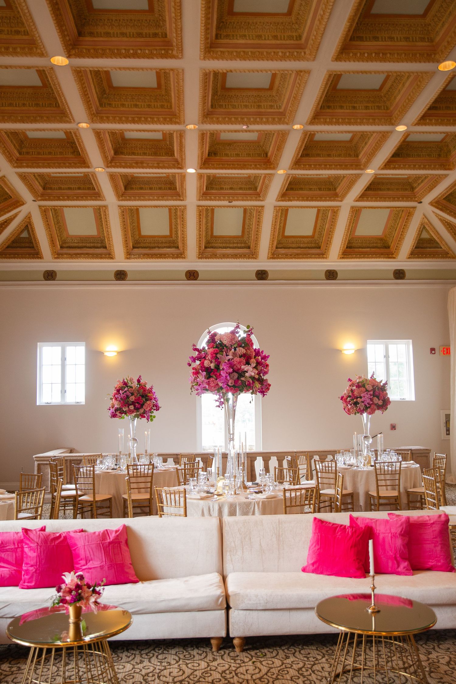 Elegant wedding reception hall with white sofas, hot pink pillows, gold tables, and tall floral centerpieces under ornate coffered ceiling.