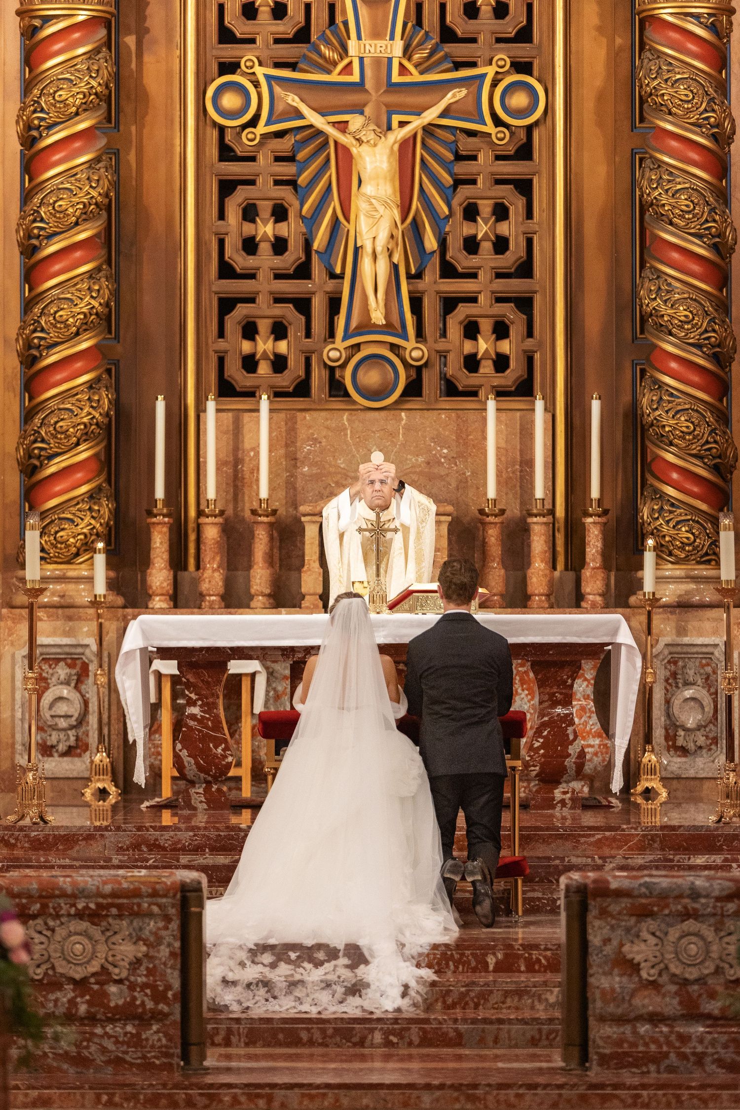 Bride and groom kneeling at church altar before priest during Catholic wedding ceremony with ornate golden crucifix.