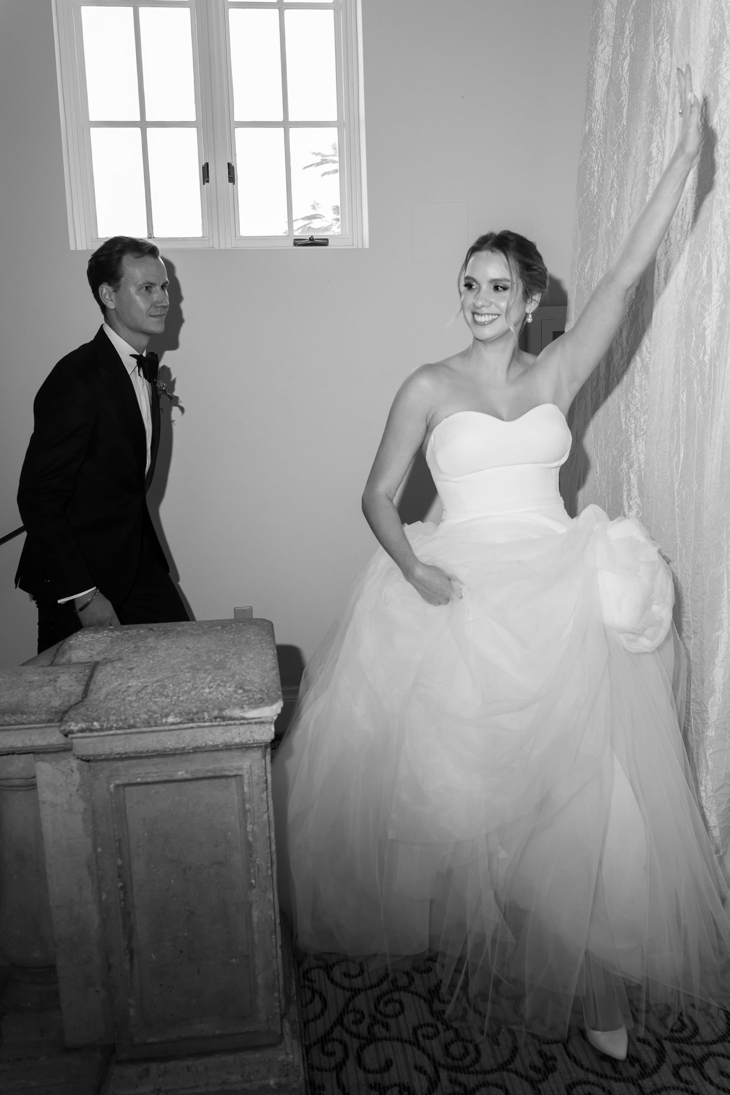 Black and white photo of a smiling bride in a strapless gown and groom in tuxedo on a staircase.