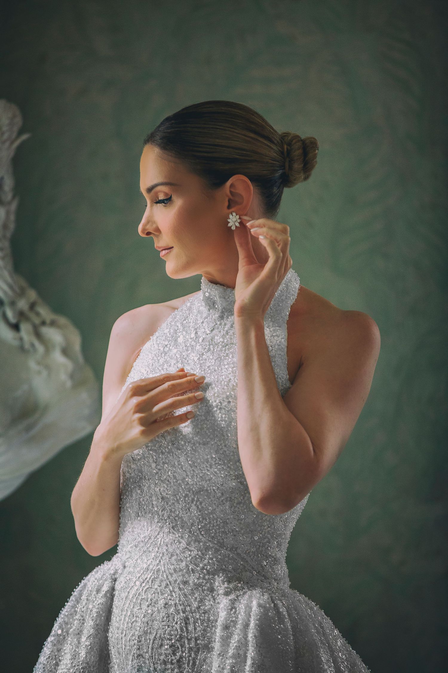 Elegant model in sparkling silver gown poses against green backdrop while adjusting earring.