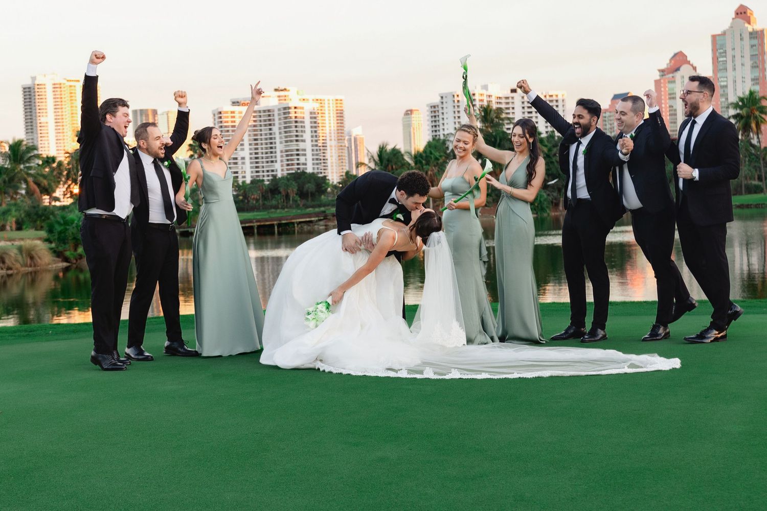 Bride and groom share a romantic dip kiss surrounded by cheering wedding party on lush green golf course with city skyline backdrop.