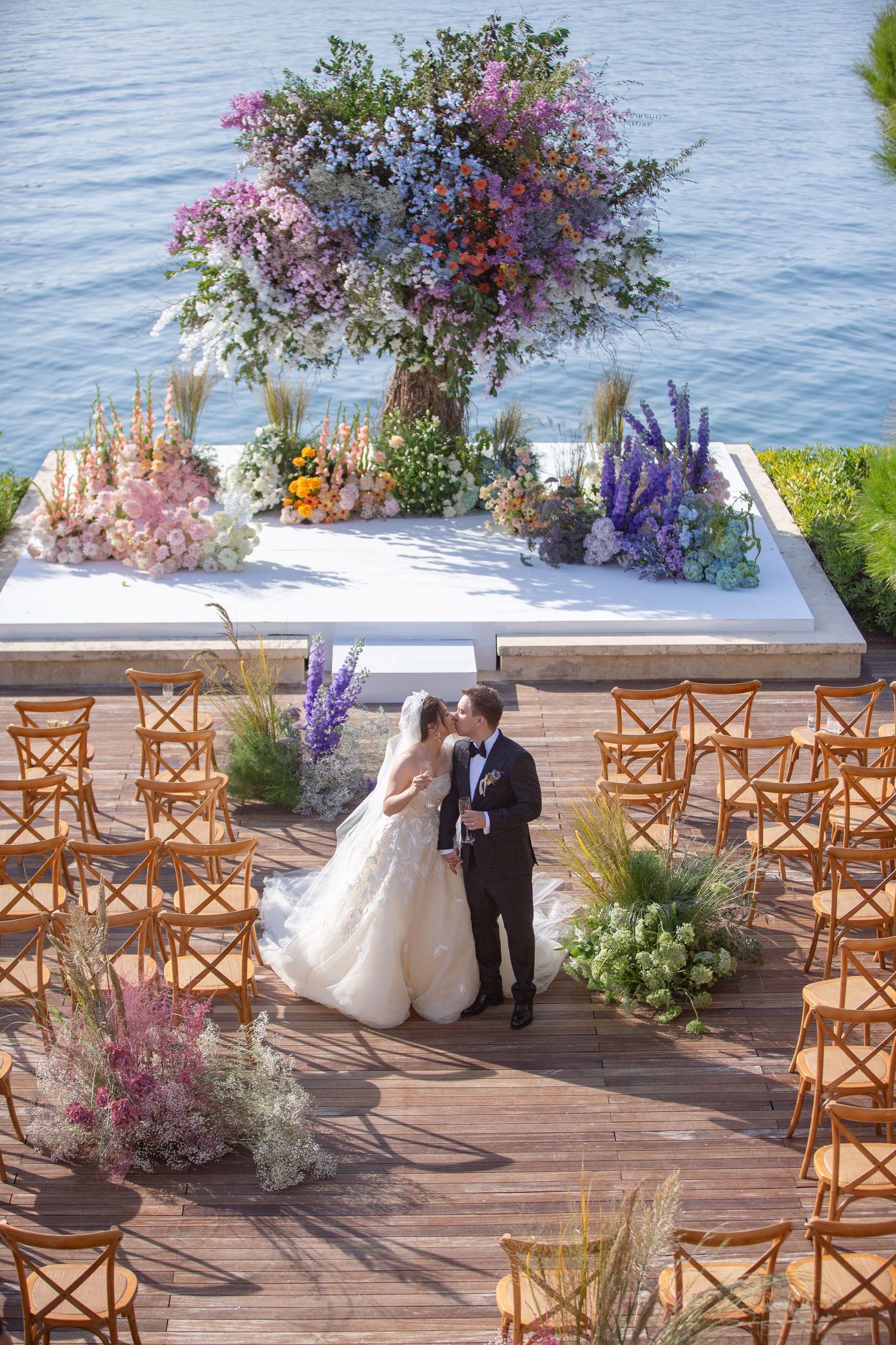 Bride and groom share a kiss at an elegant outdoor waterfront wedding ceremony with colorful floral arrangements.