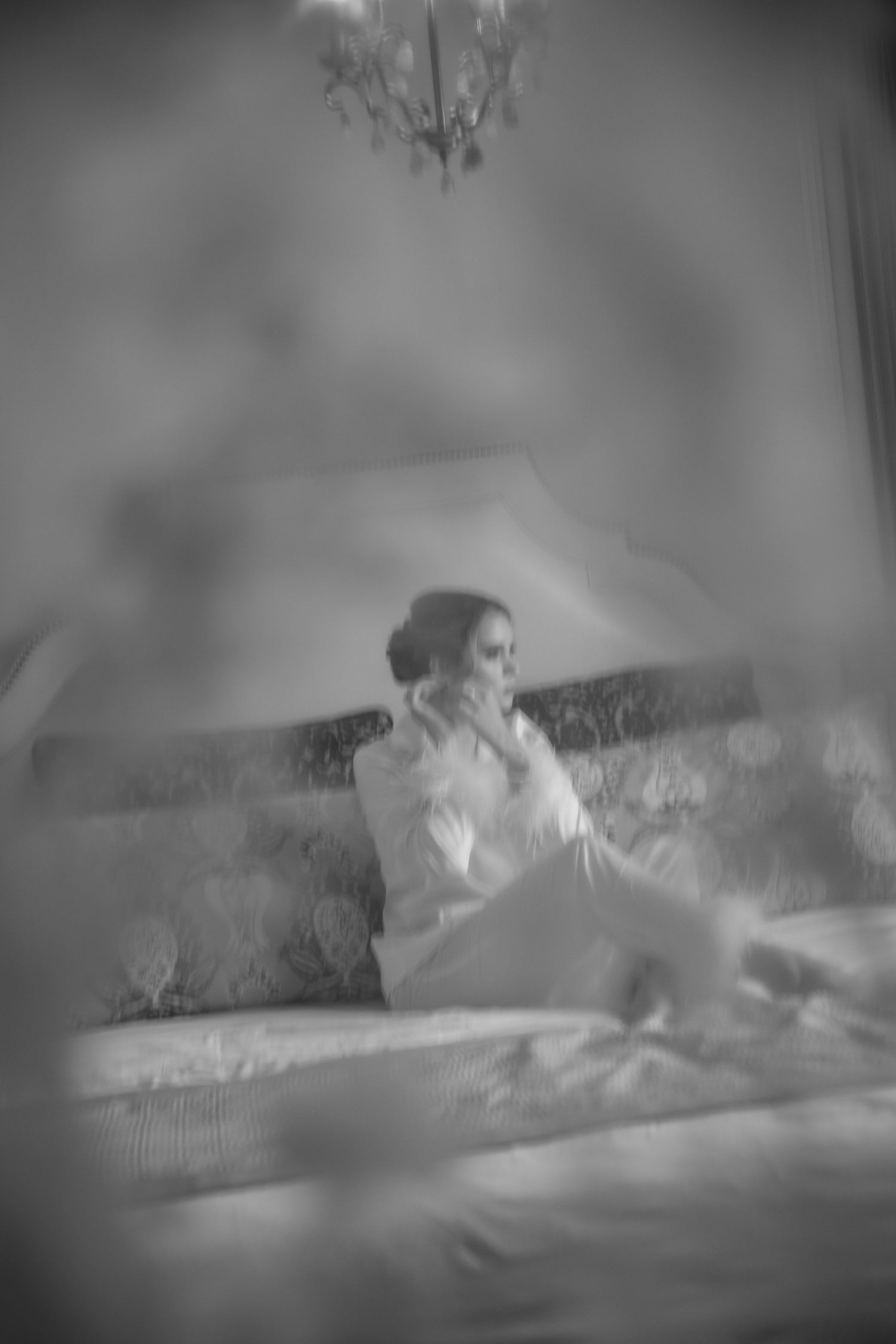 Black and white photo of a bride in white robe sitting on bed, reflected in mirror with soft dreamy bokeh effect.