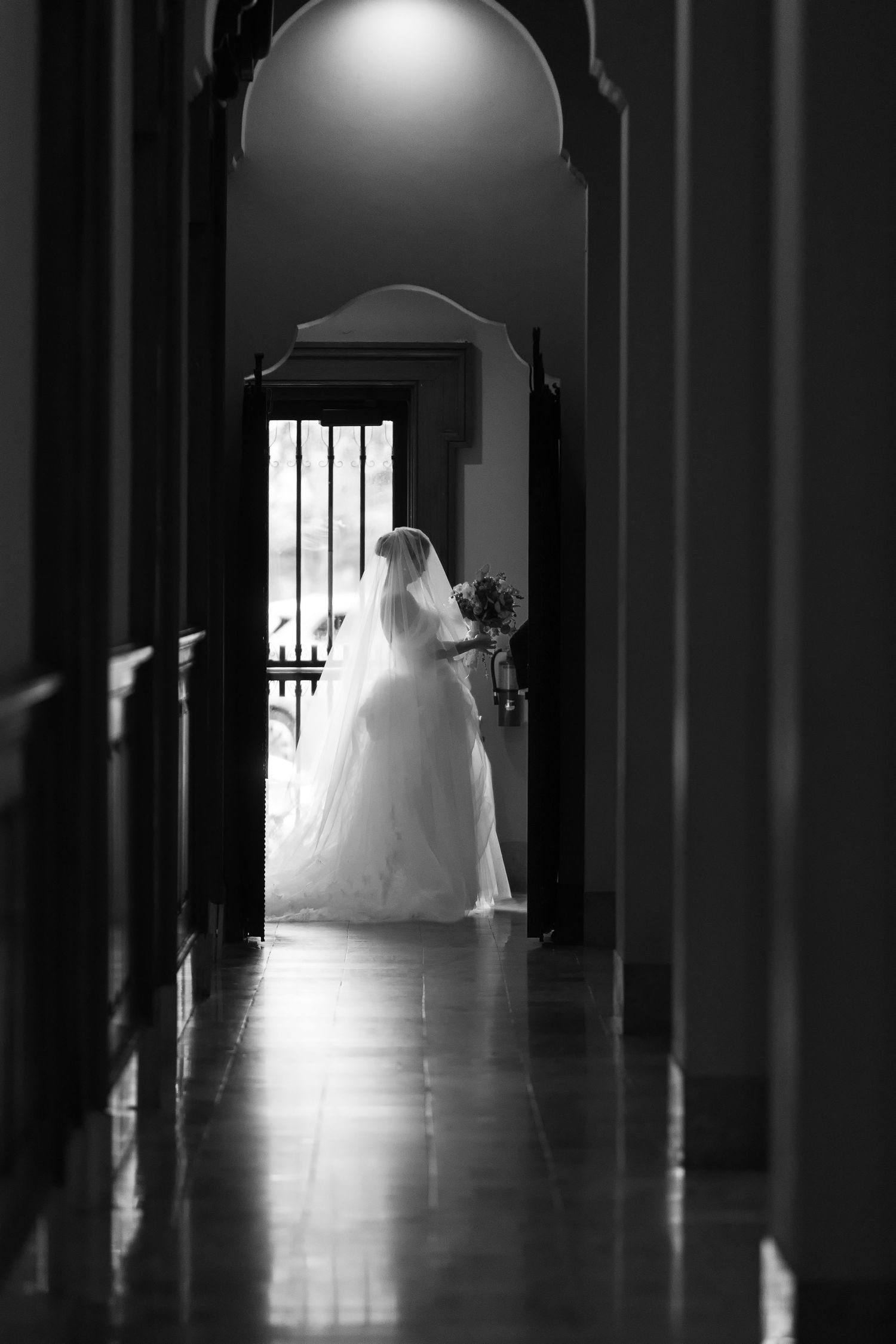 Black and white photo of a bride in a flowing gown and veil holding a bouquet, standing in a sunlit doorway.