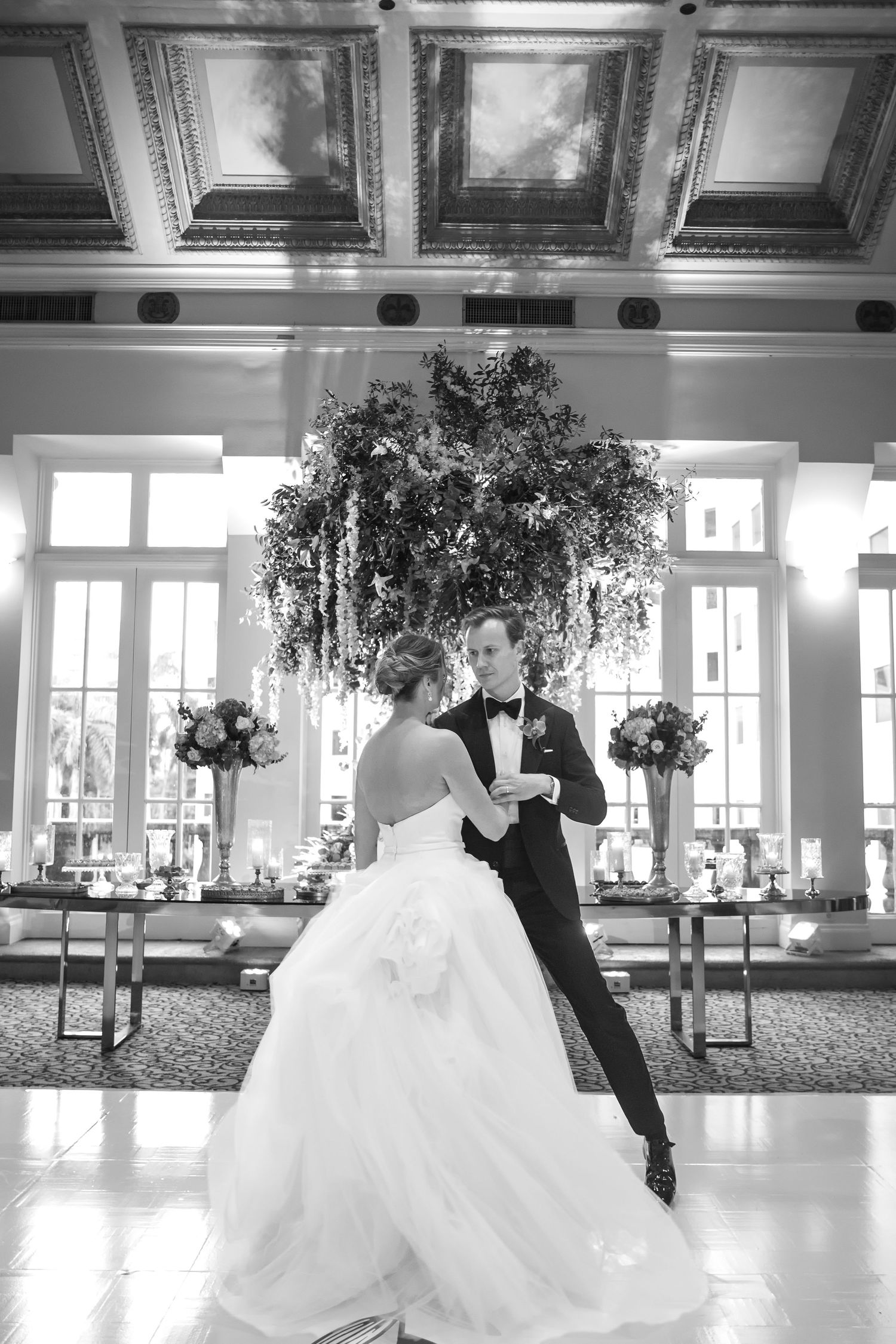 Black and white photo of bride and groom sharing first dance in elegant ballroom with floral centerpiece.
